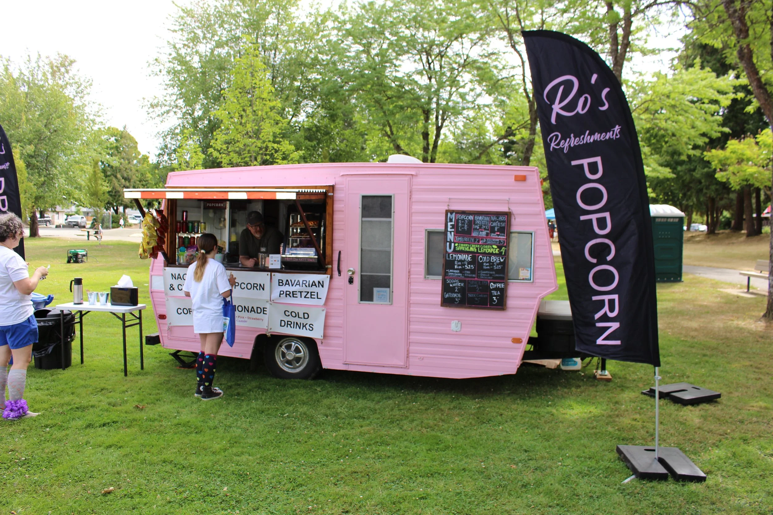 Pink food truck selling popcorn, Bavarian pretzels, and cold drinks at an outdoor event, with two people waiting for orders and a black flag that reads 'Ro's Refreshments POPCORN'