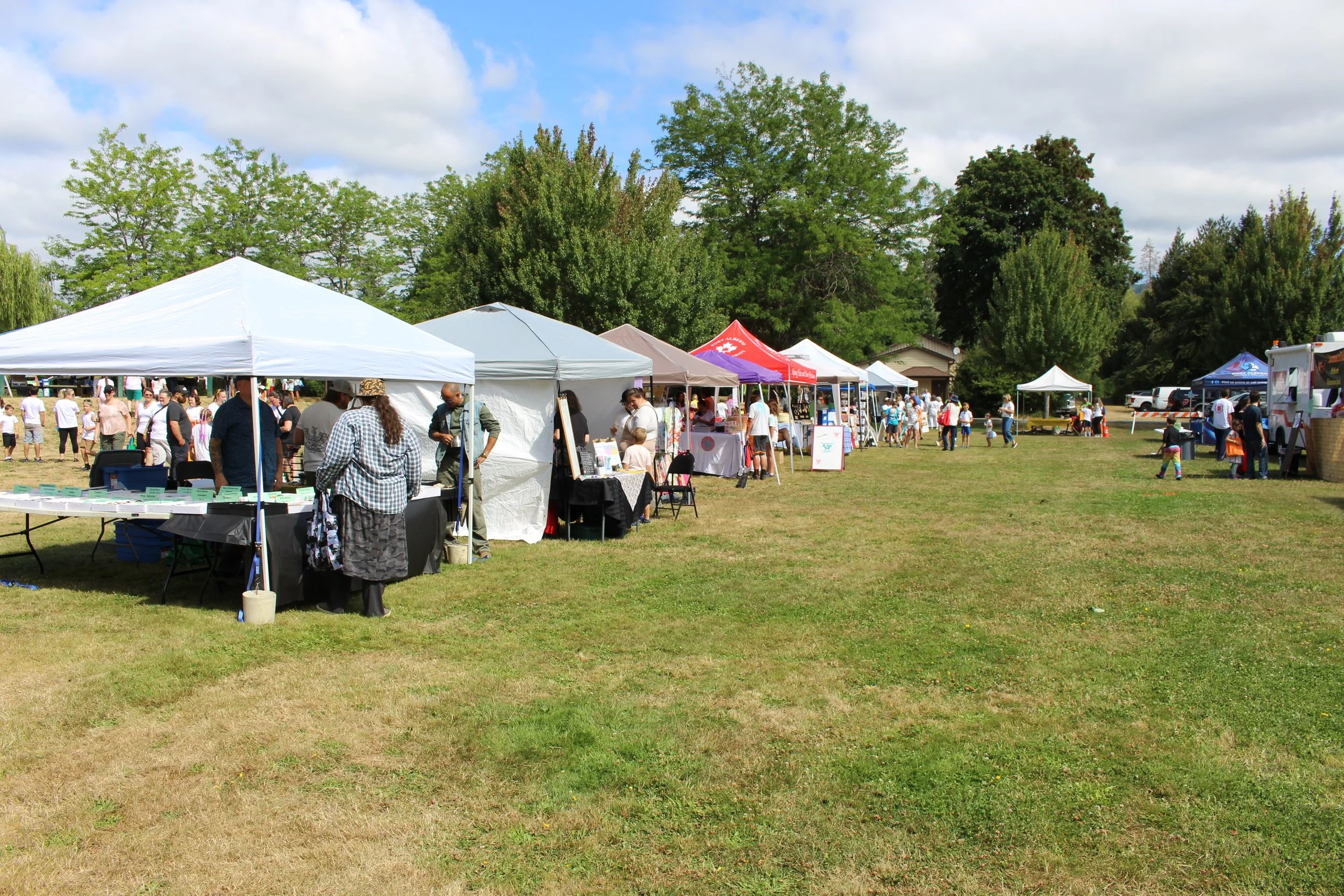 An outdoor fair with multiple vendor tents set up on a grassy field under a partly cloudy sky, with people browsing and walking around.