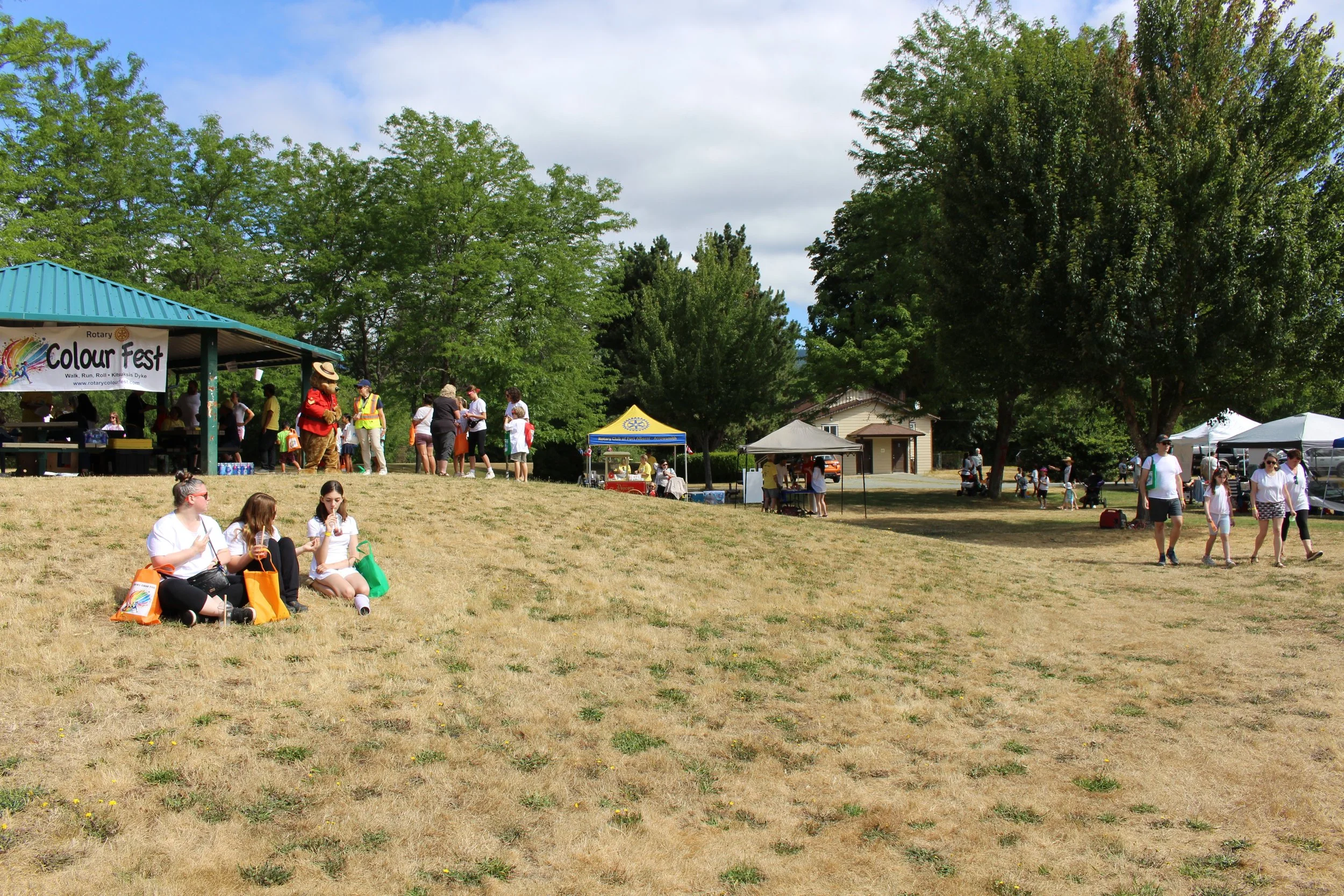 People gathering at a community outdoor event called 'Colour Fest,' with tents, a stage, and a grassy area with trees, some sitting and some walking.