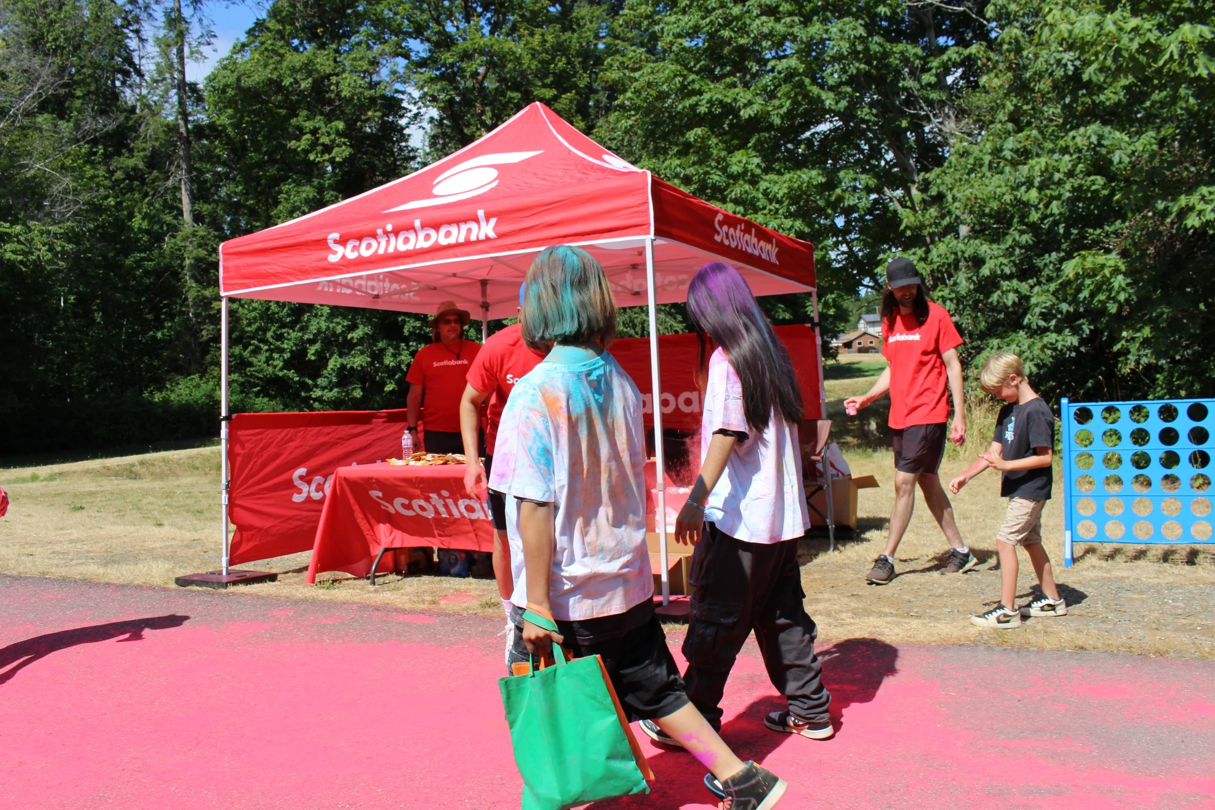 People participating in a color run event near a red Scotiabank tent, with colorful powder on their clothes, walking on pink-colored ground surrounded by trees.