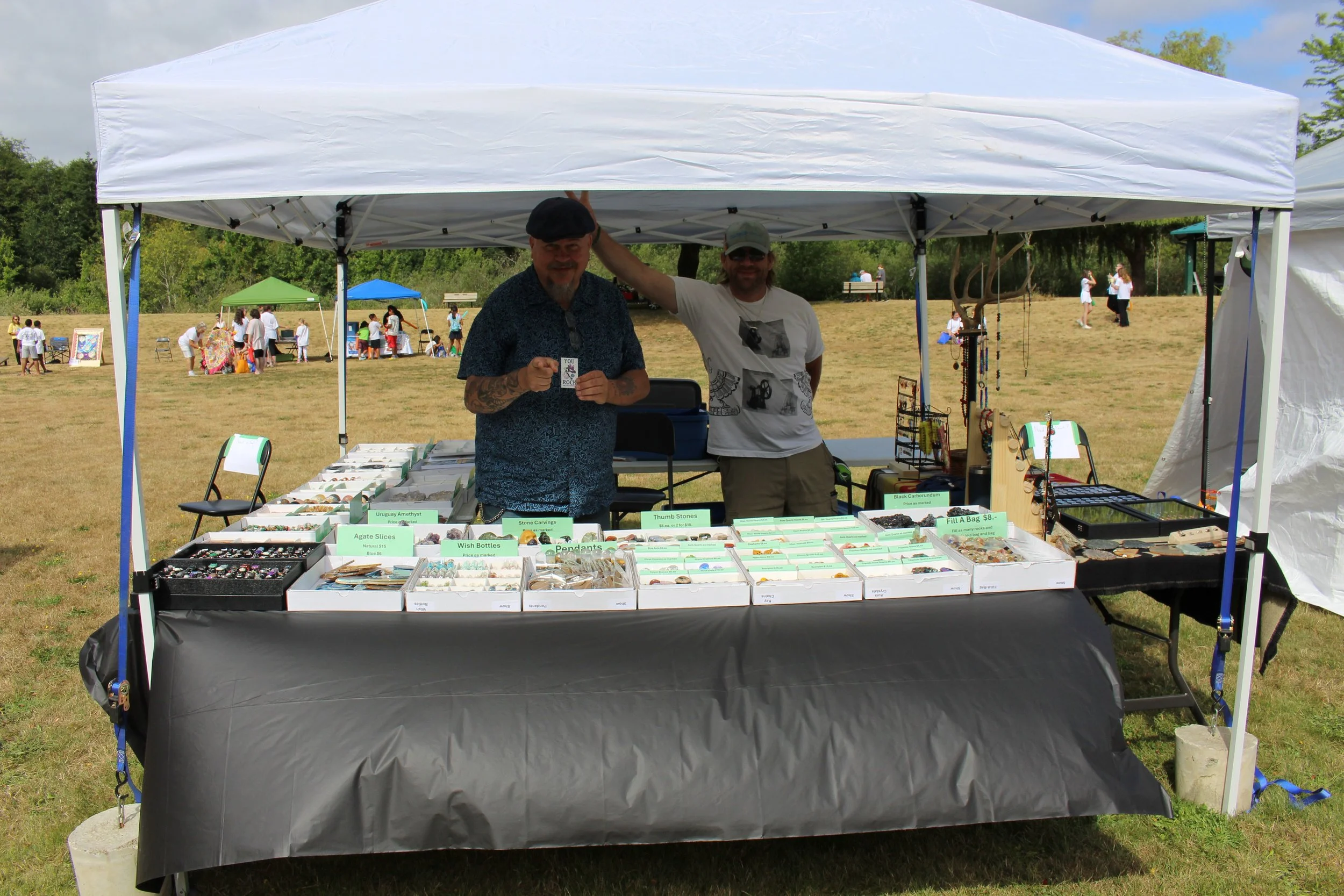 Two men standing behind a table at an outdoor market booth selling jewelry and various trinkets under a white canopy tent.