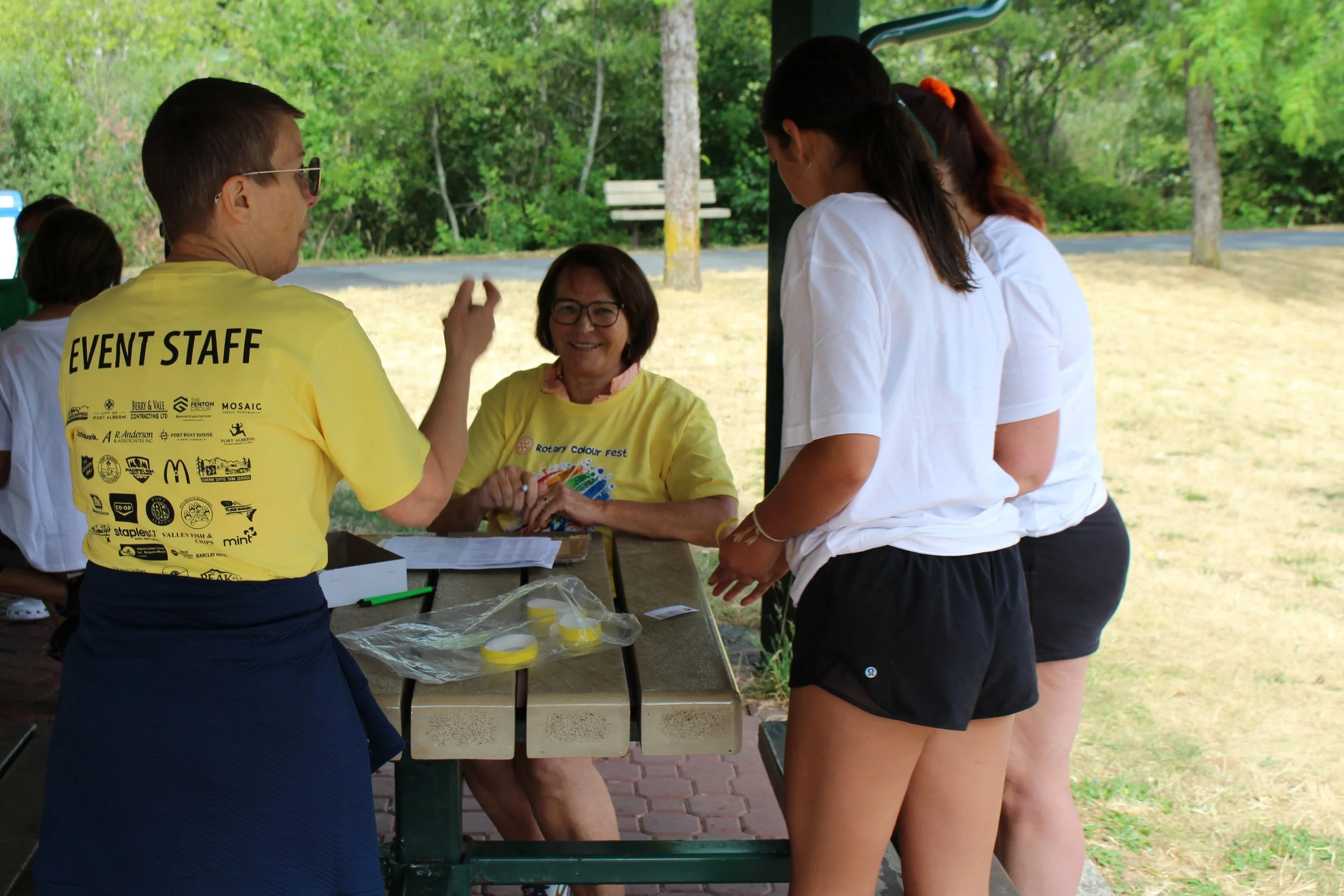 Four women talking at an outdoor registration or check-in table, with trees and grass in the background. Two women are wearing yellow event staff shirts, and the other two women are wearing white shirts and black shorts.