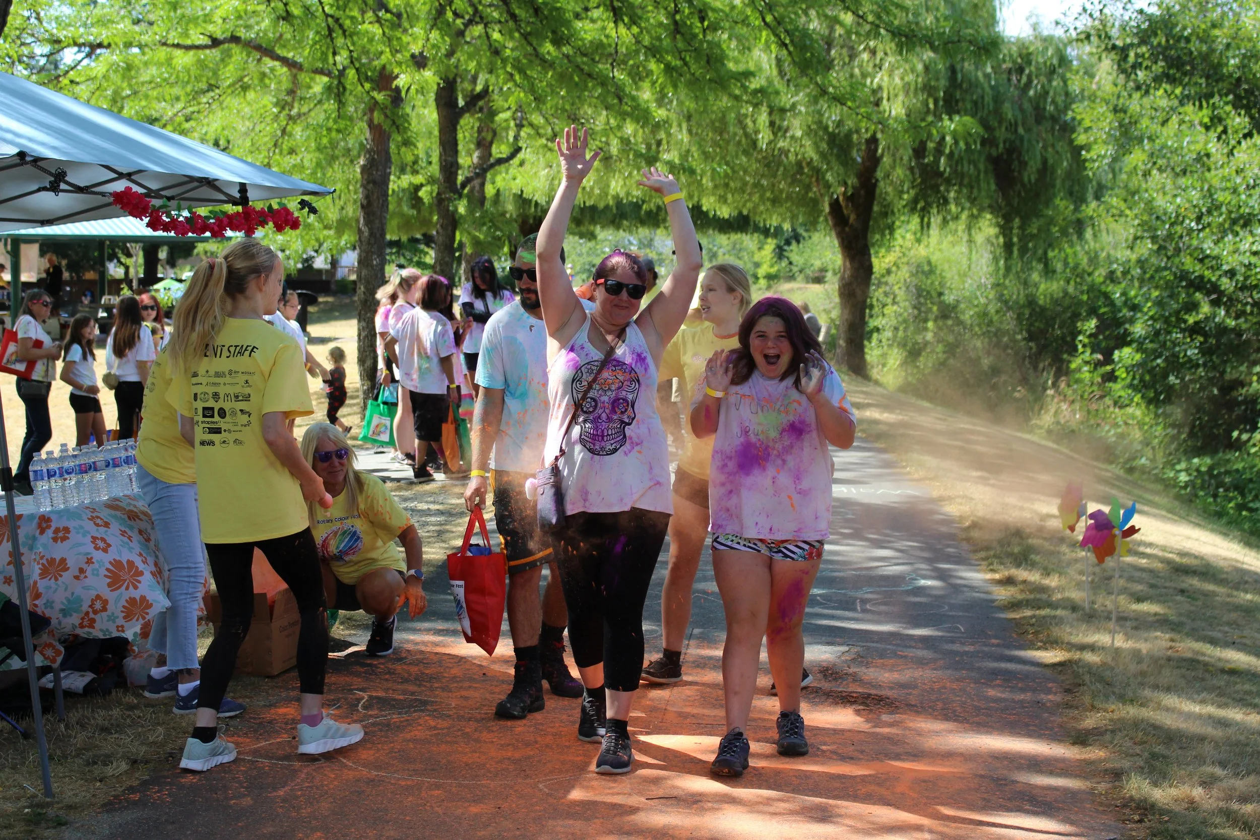 People participating in a colorful outdoor event, throwing colored powder, with trees and a booth in the background.