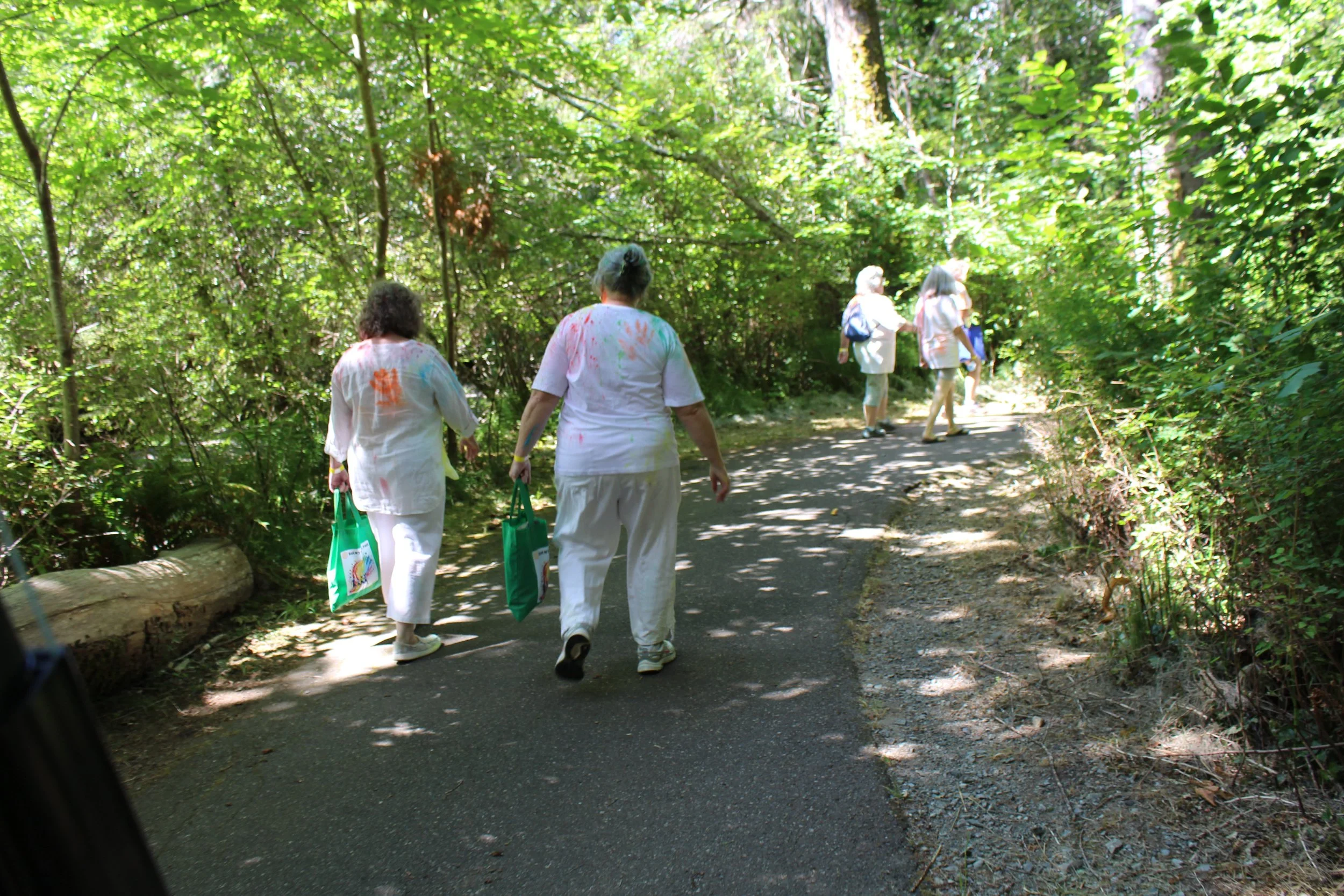 Group of people walking on a shaded forest trail, some wearing white clothing with colorful paint stains, carrying green bags.