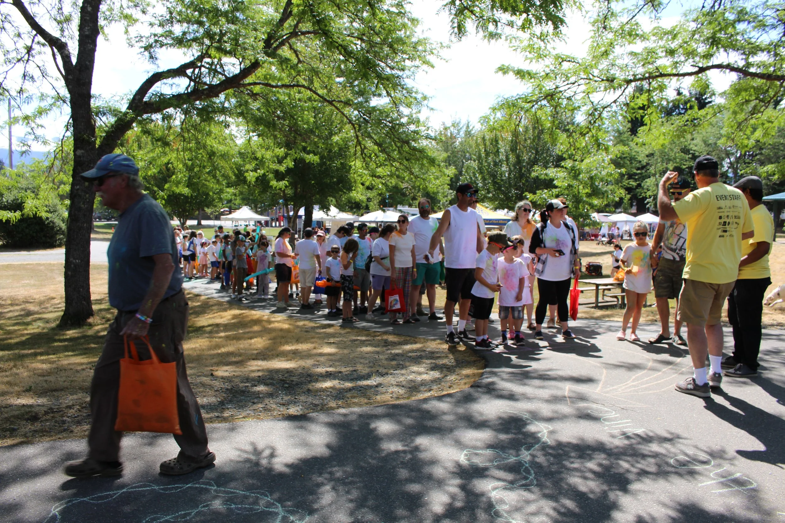 Children and adults in a line outdoors at a park during daytime, with trees and tents in the background.