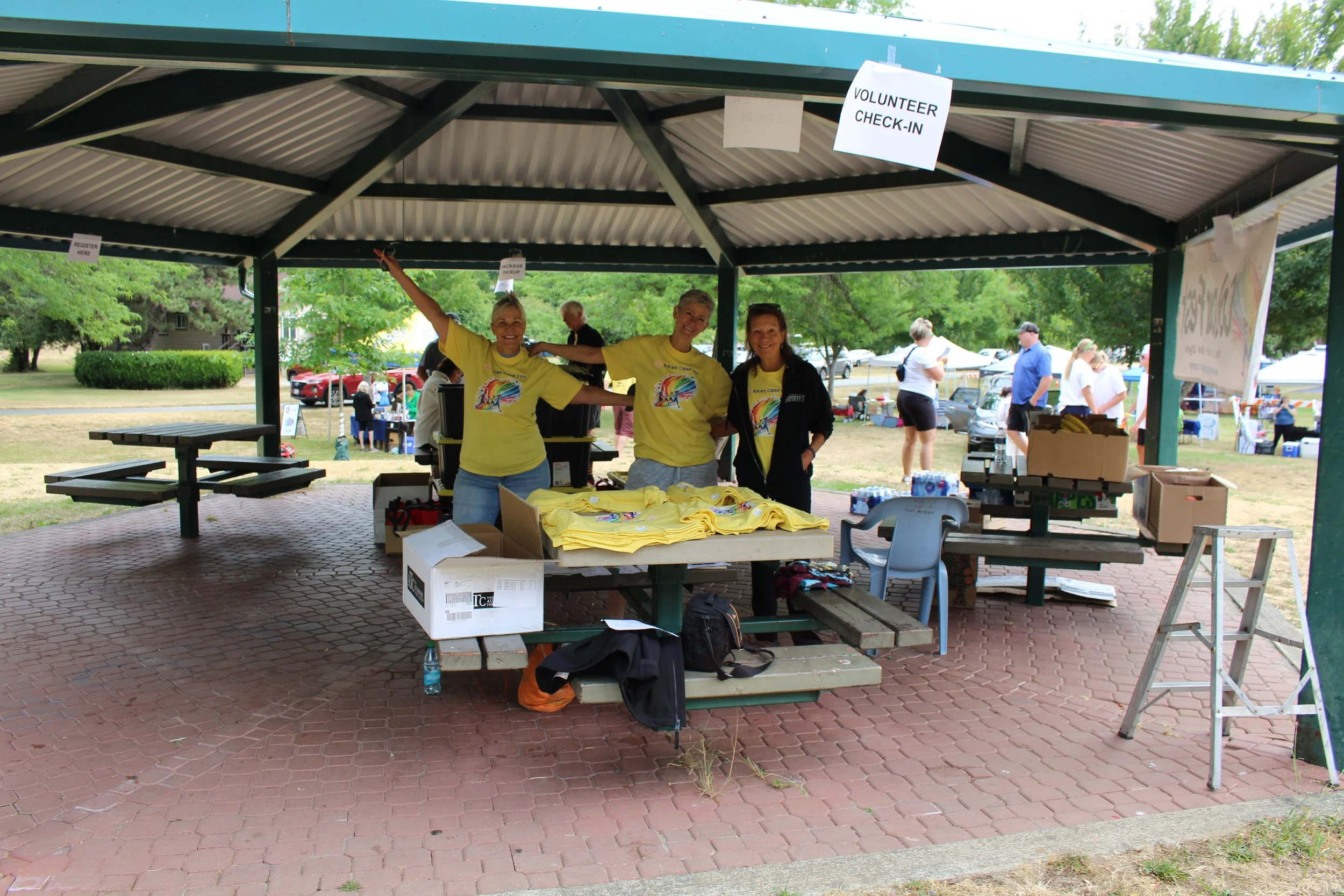 Three volunteers in yellow shirts standing under a pavilion with a sign that says 'Volunteer Check-In,' at an outdoor event. They are smiling and posing for the photo, with tables of yellow shirts and supplies in front of them.