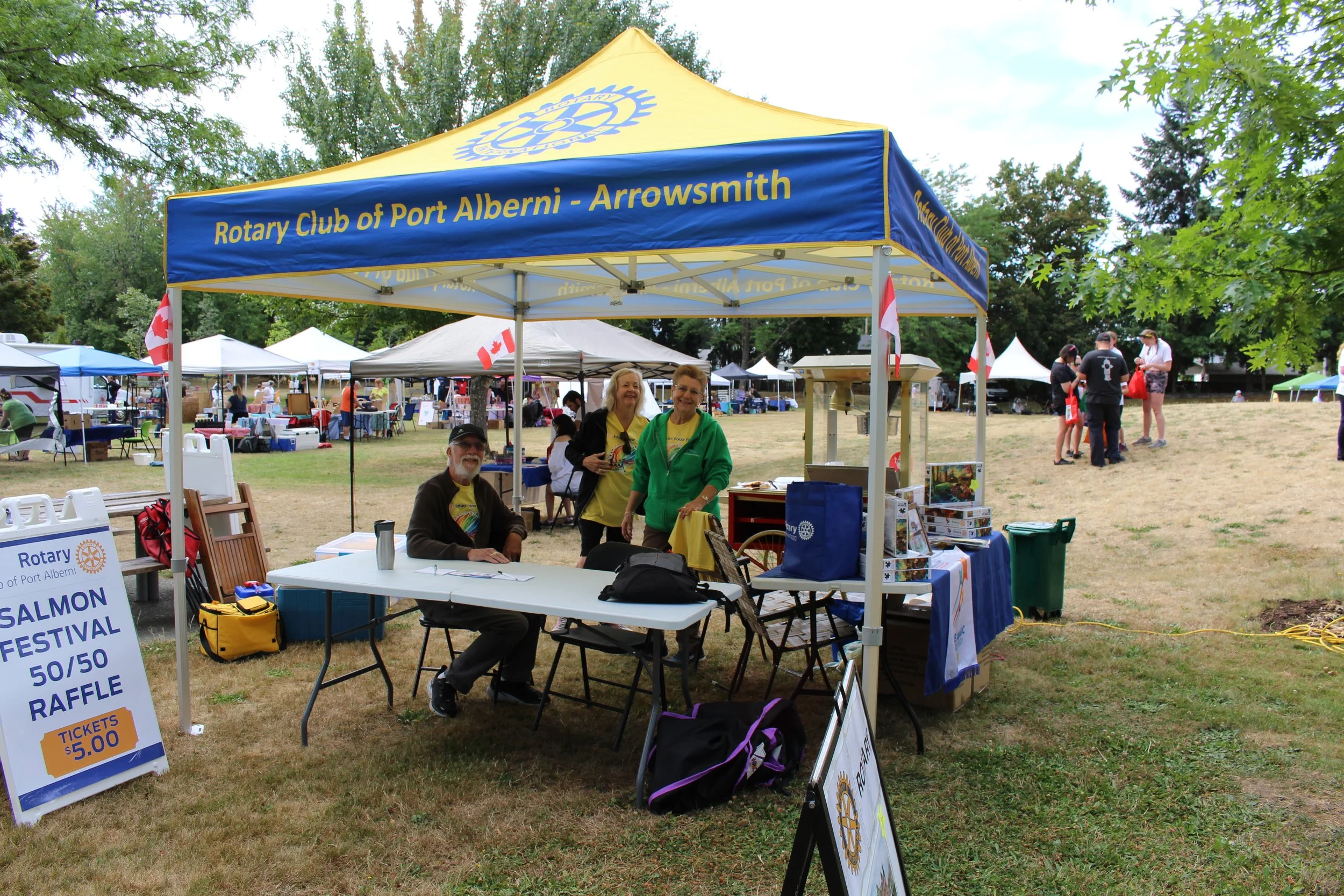 A Rotary Club booth at an outdoor festival with a yellow and blue canopy. The booth has a sign promoting a salmon festival raffle. People are sitting and standing around the booth, and other tents are visible in the background at the outdoor event.