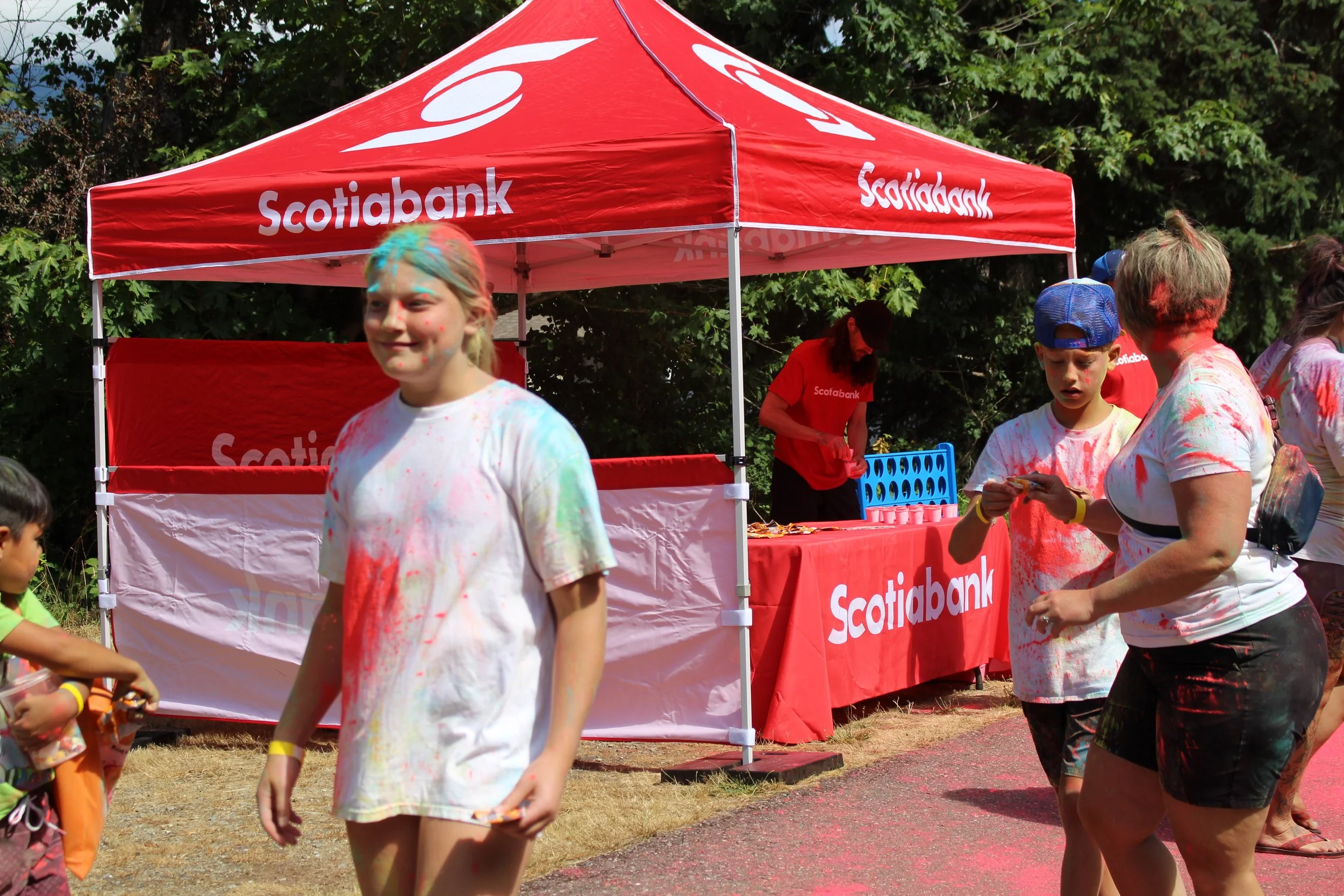 People participating in a color run event, with a red Scotiabank tent in the background, splattered with colored powder on their clothes and faces.