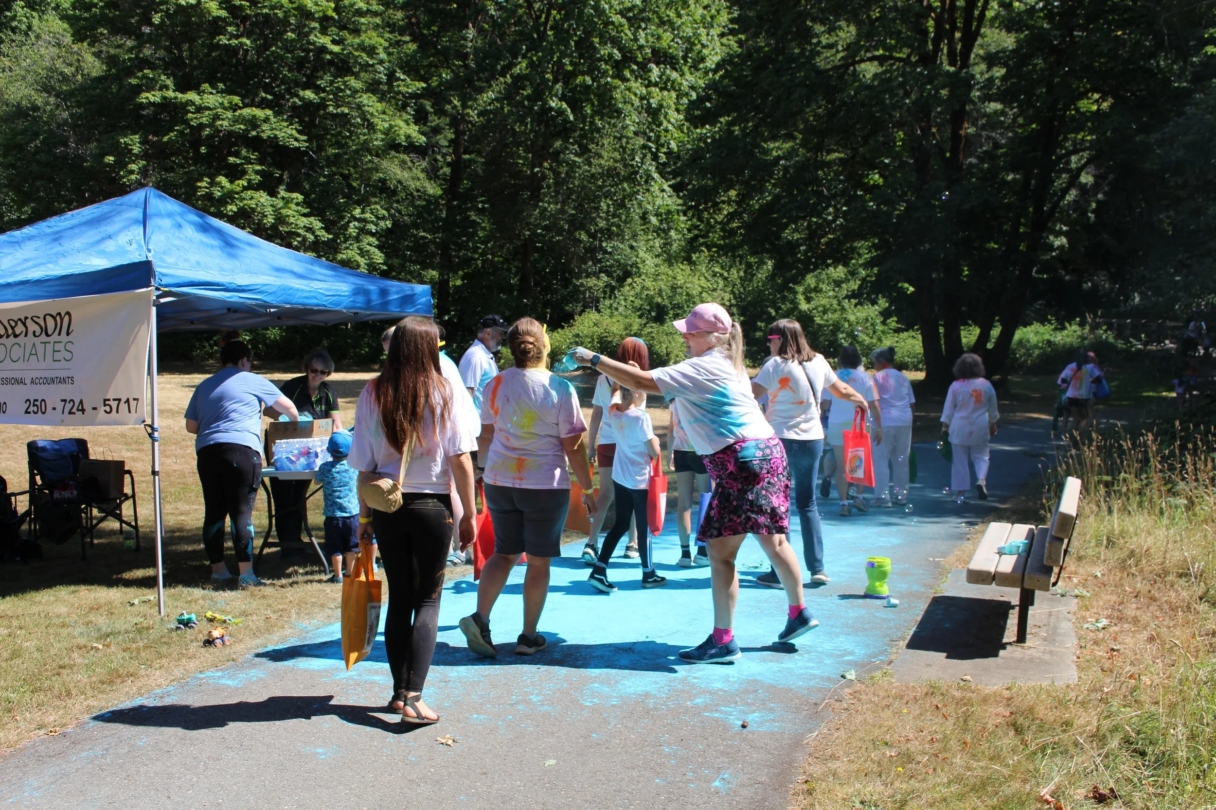 People participating in a color run event outdoors, with some throwing colored powder, under a blue tent, surrounded by green trees.