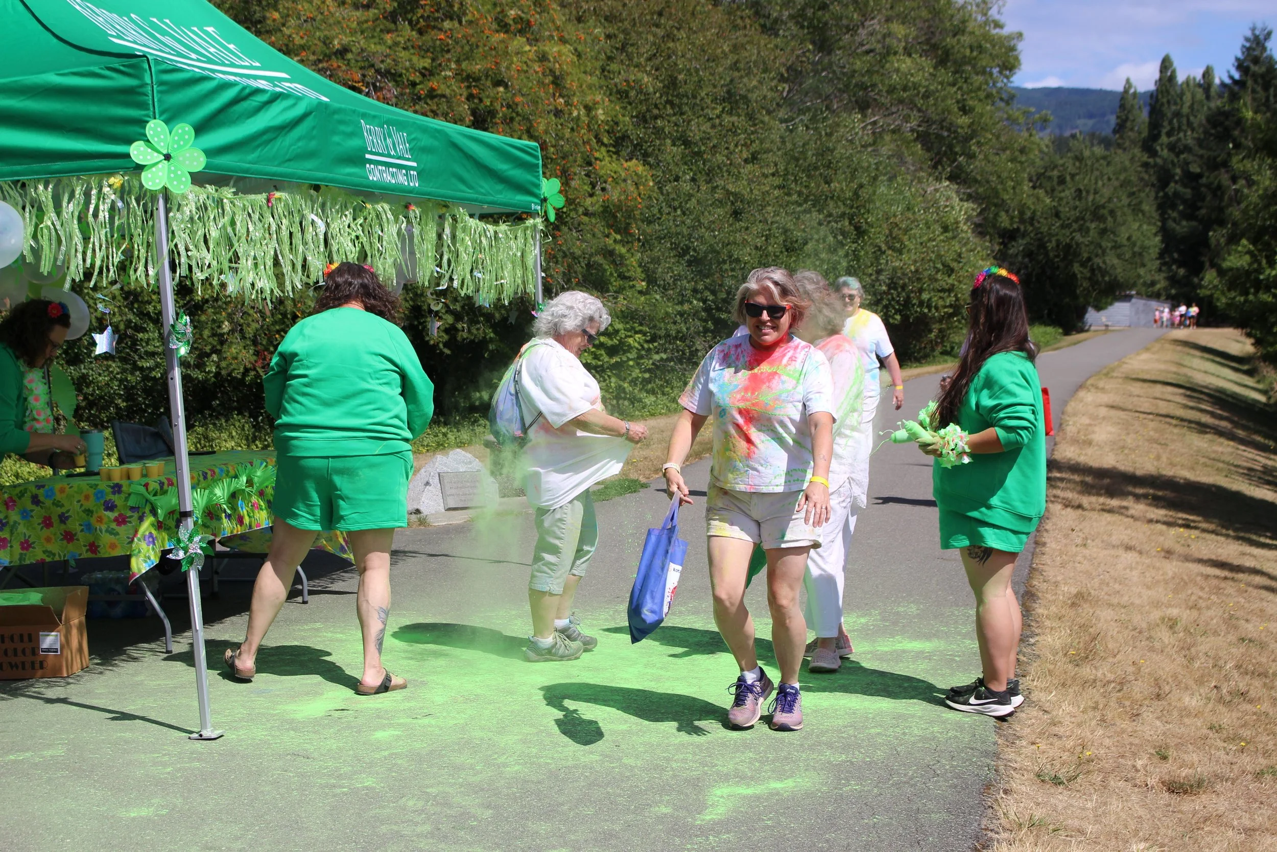 People celebrating St. Patrick's Day outdoors, with green clothing, participating in a color throw event near a green tent decorated with shamrocks and green streamers.
