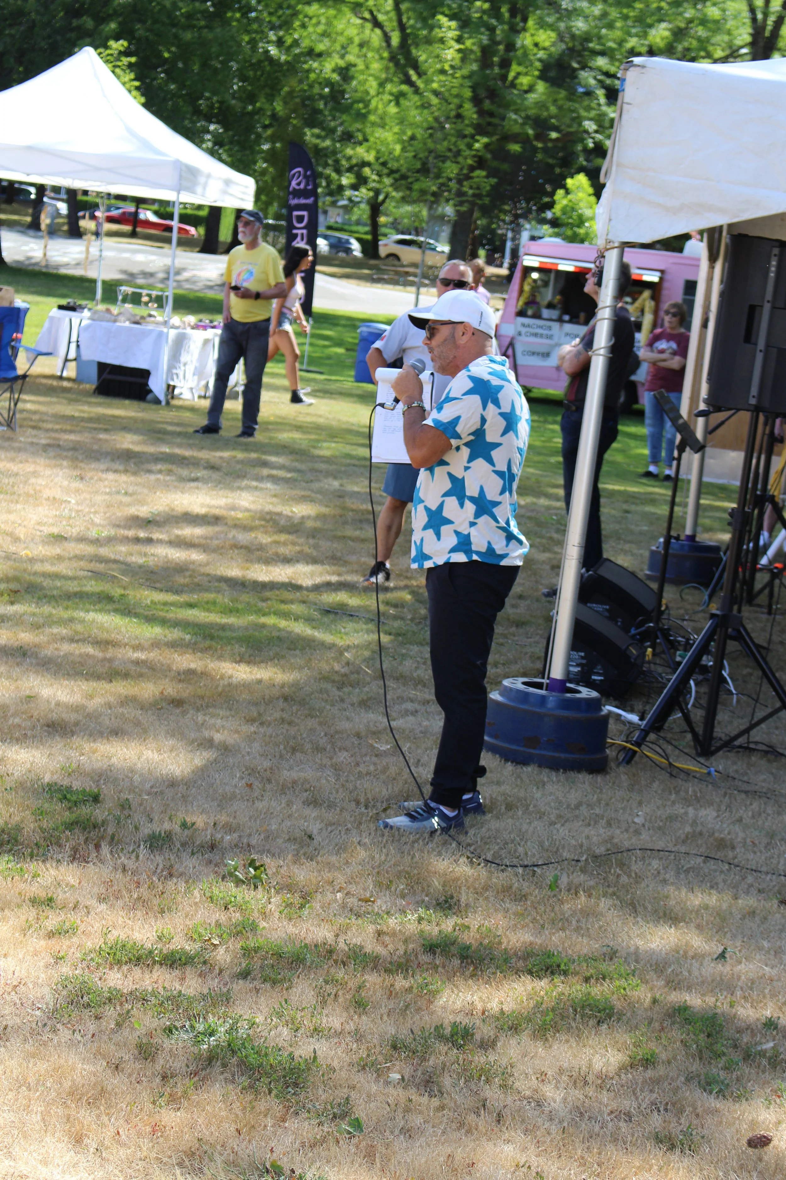 Man wearing a white cap and star-patterned shirt speaking into a microphone at an outdoor event with tents, people, and a pink food truck in the background.