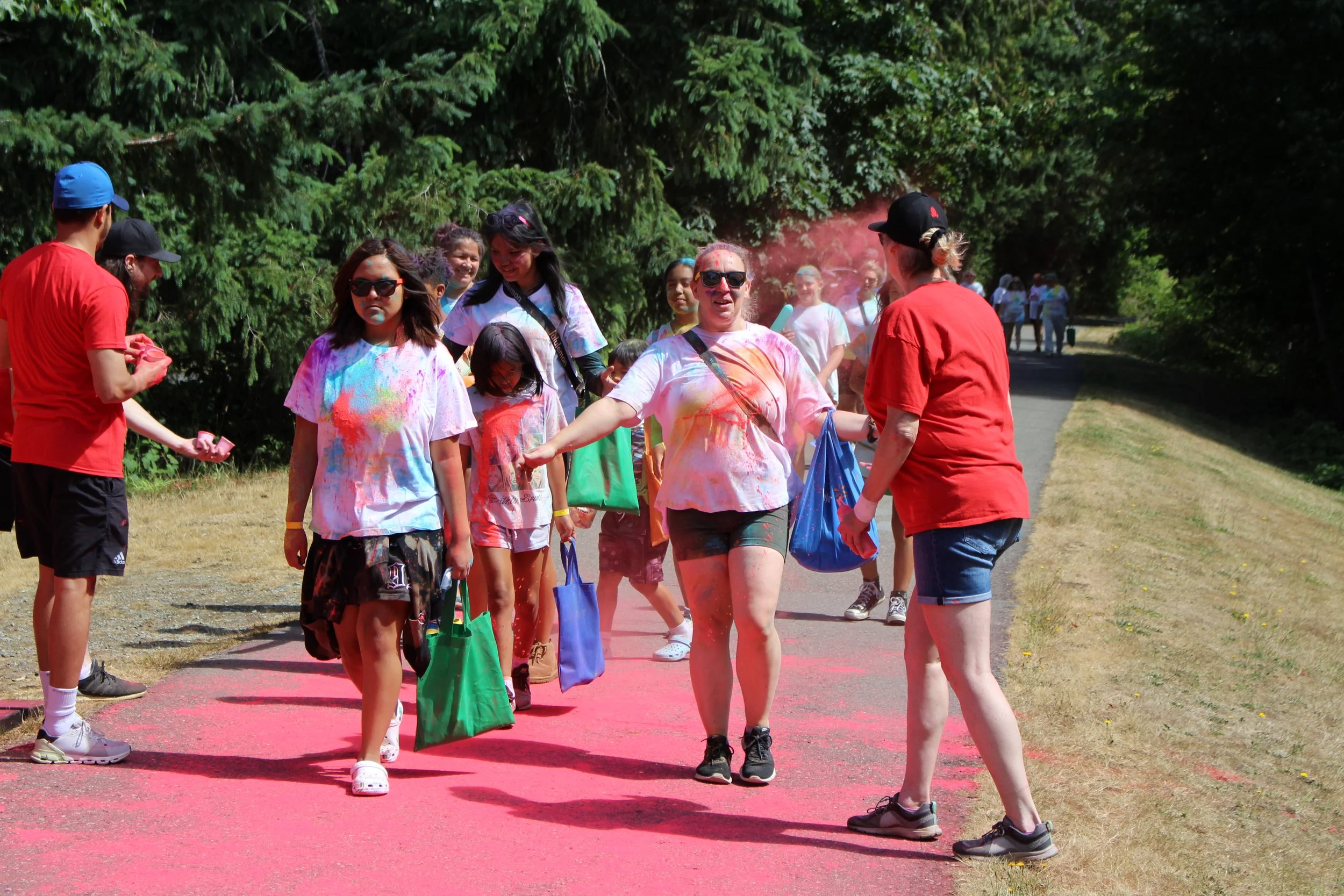 People participating in a color run, covered in colorful powder, walking along a pink path in a park with trees in the background.
