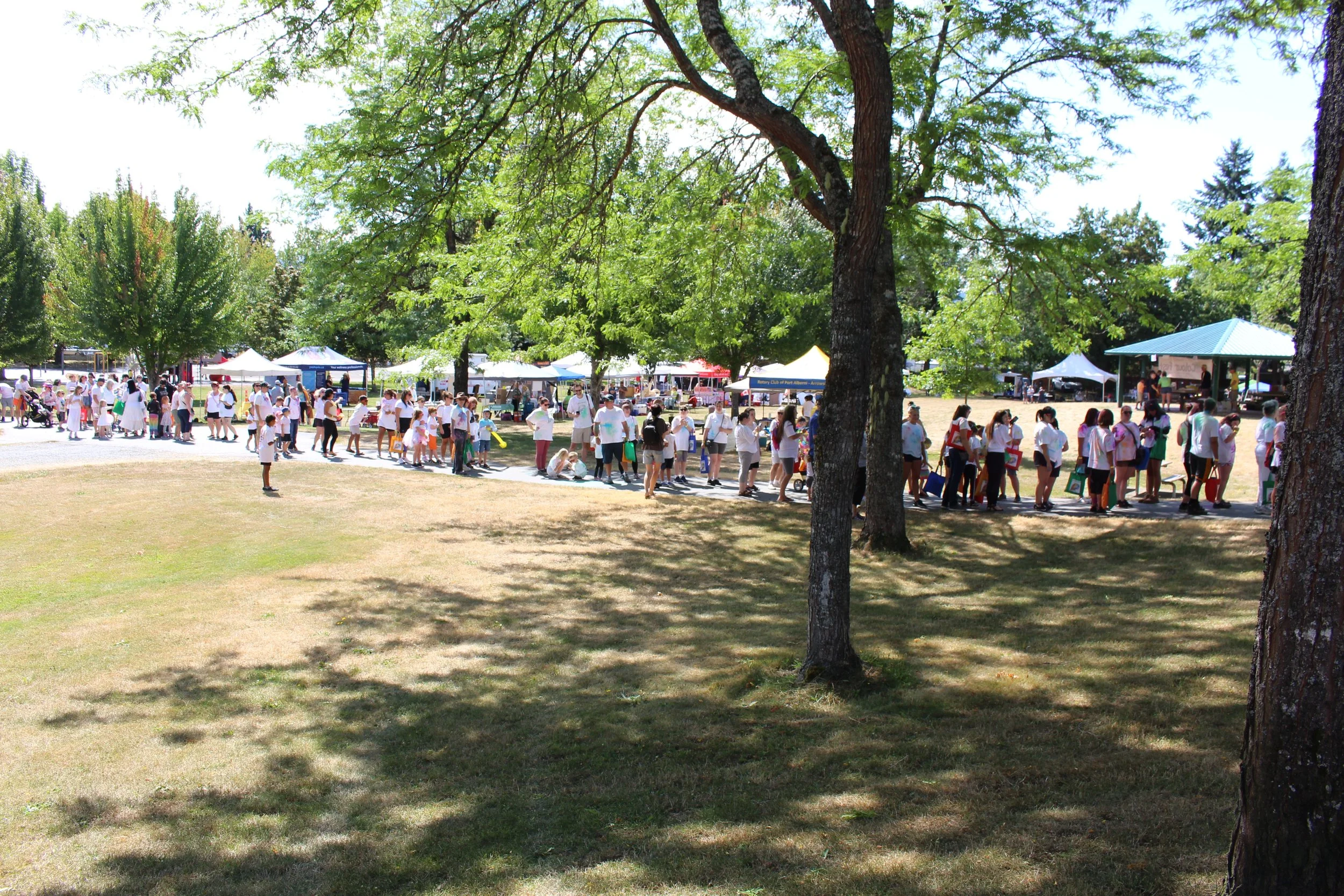 Crowd of people waiting in line outdoors at a sunny park with trees and tents.