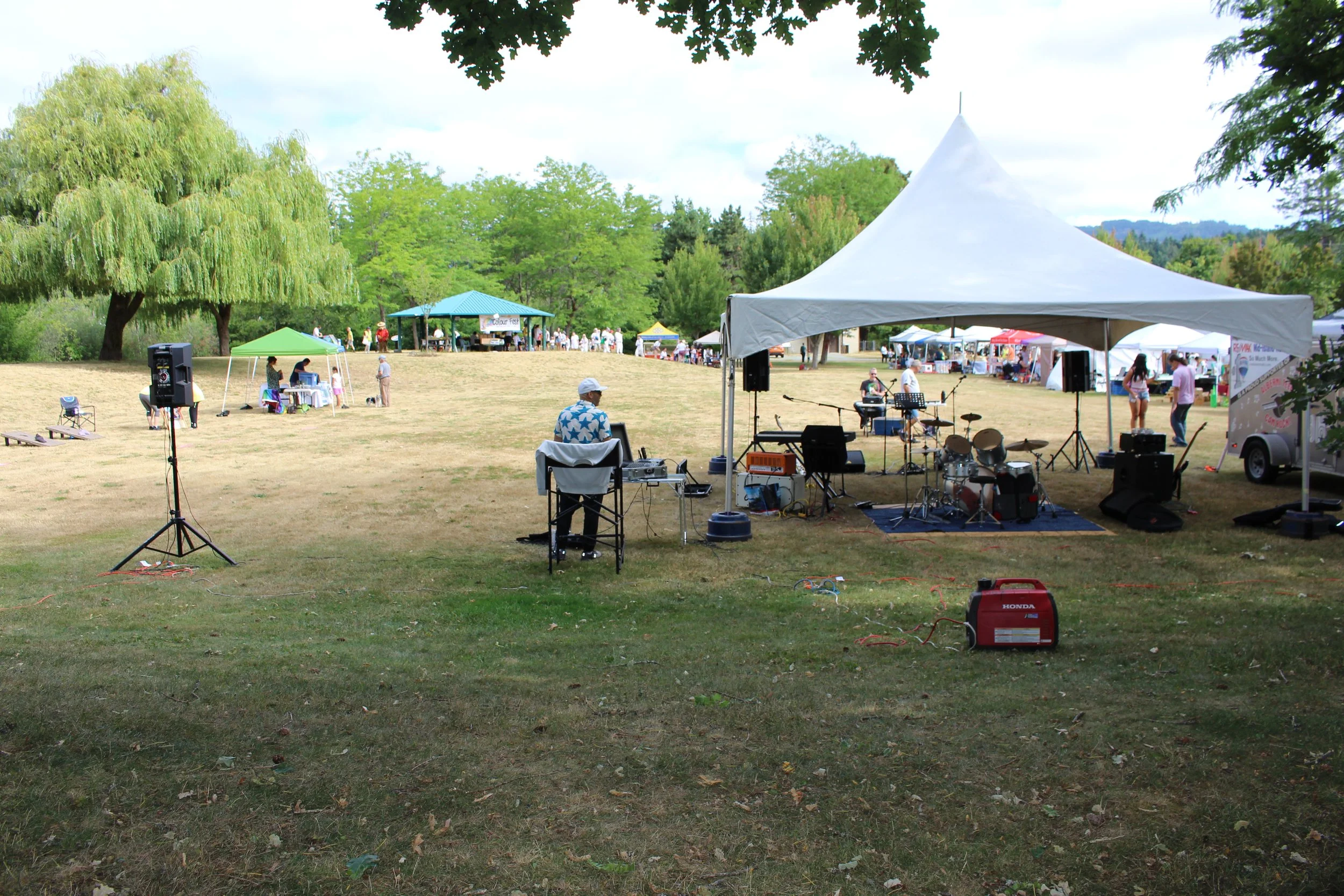 An outdoor stage with musical instruments under a large white canopy, set up in a park with people, tents, and trees in the background during a festival or outdoor event.