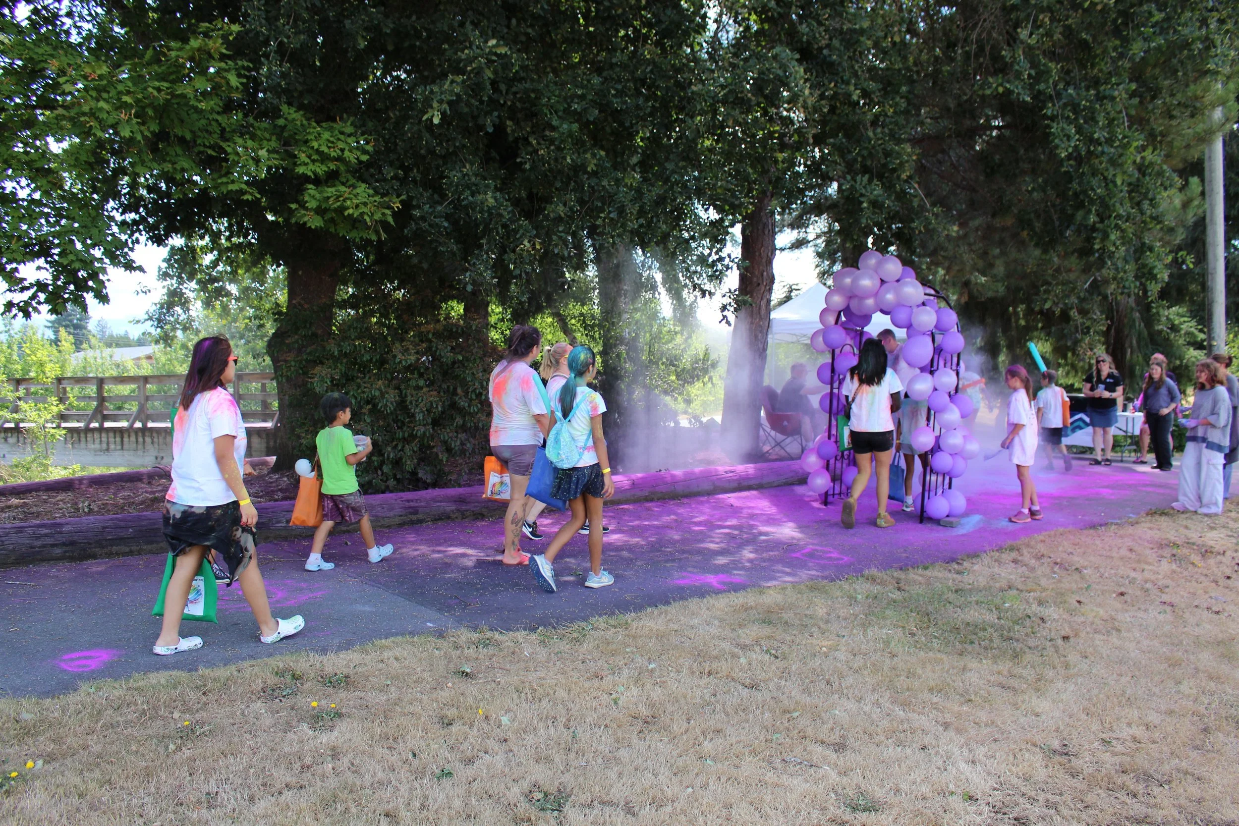 People participating in a color run event, walking through a purple powder station with a balloon arch entrance, outdoors on a sunny day surrounded by trees.