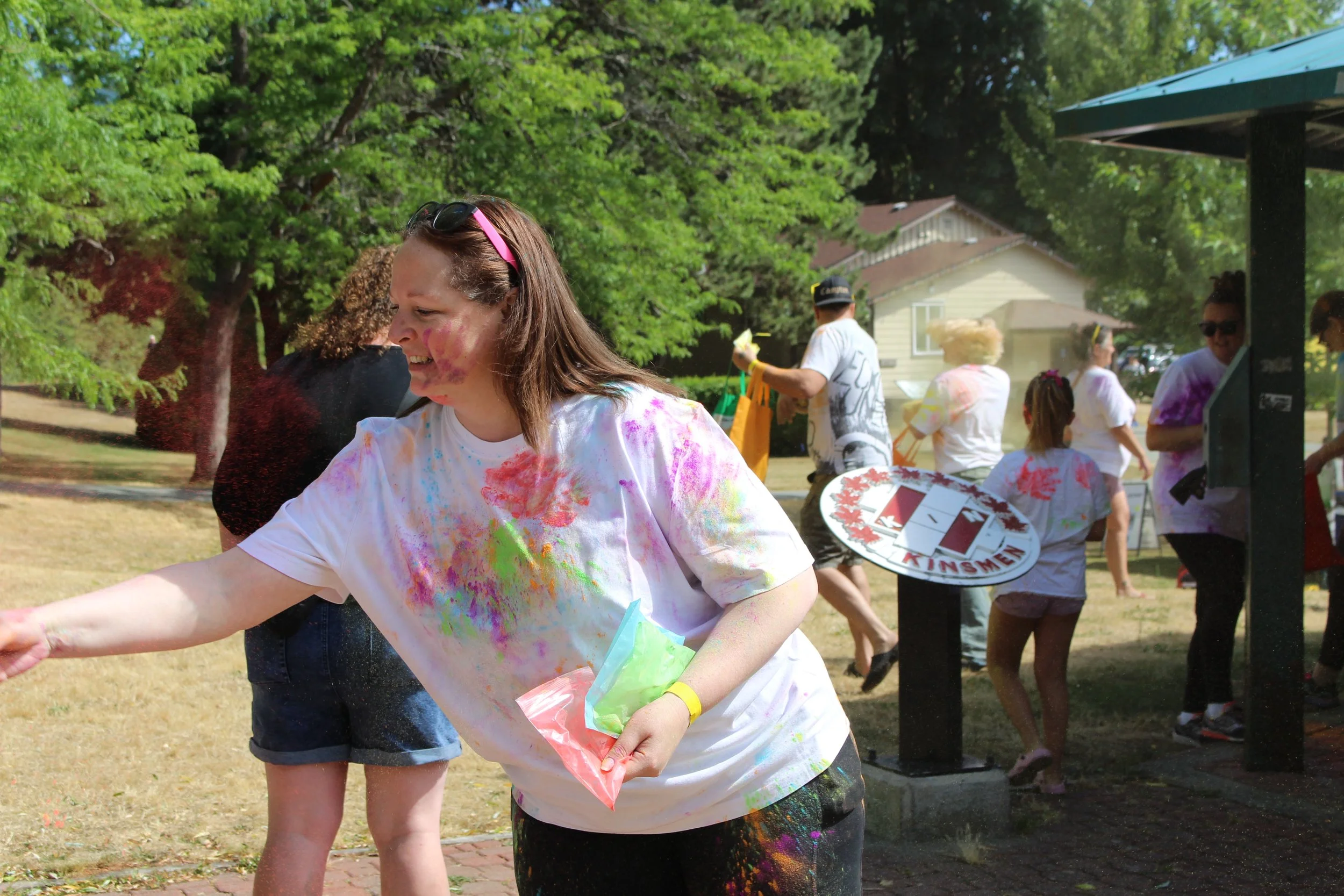 People celebrating at a color run event, with a woman smiling and covered in colorful powder, others in the background are also covered in color, some are holding color packets and there is a sign with the words "Kinsmen".