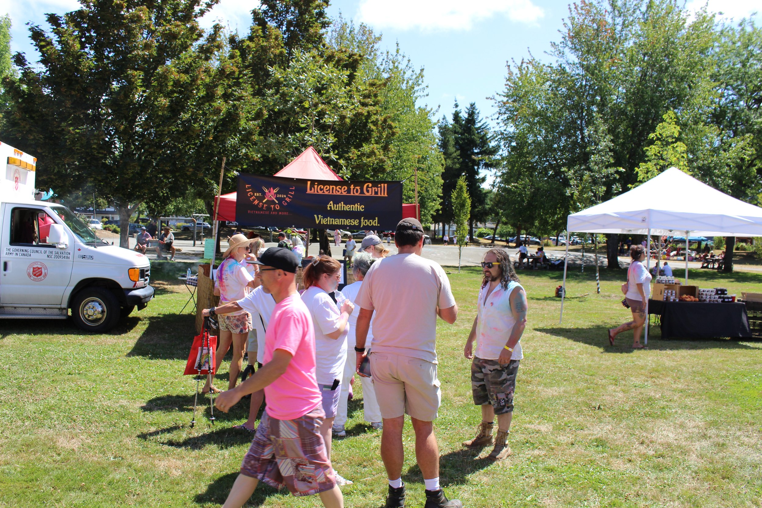 People gather around a food truck at an outdoor festival, with a sign reading 'License to Grill' and offering authentic Vietnamese food. There are trees and additional vendor tents in the background.