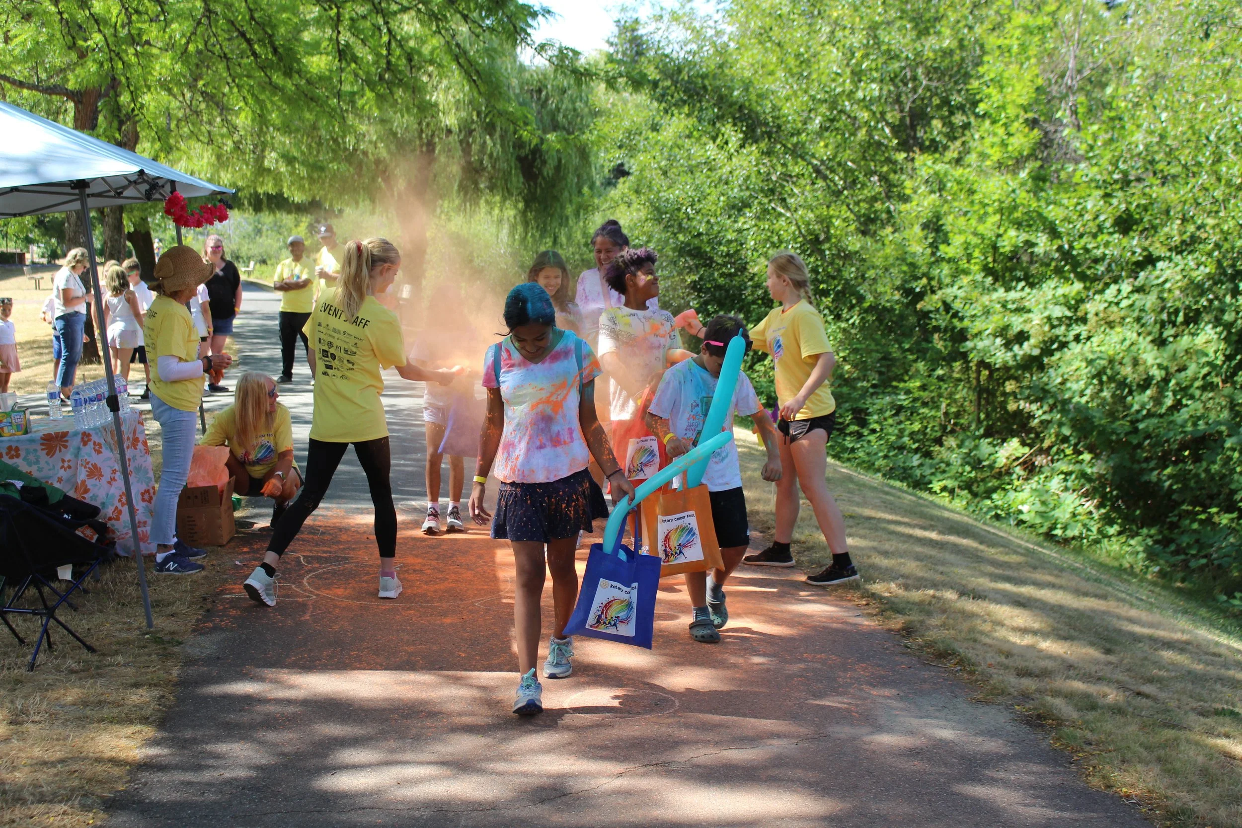 Children and adults celebrating at a color run event outdoors, with some children covered in colored powder, on a shaded path lined with trees.