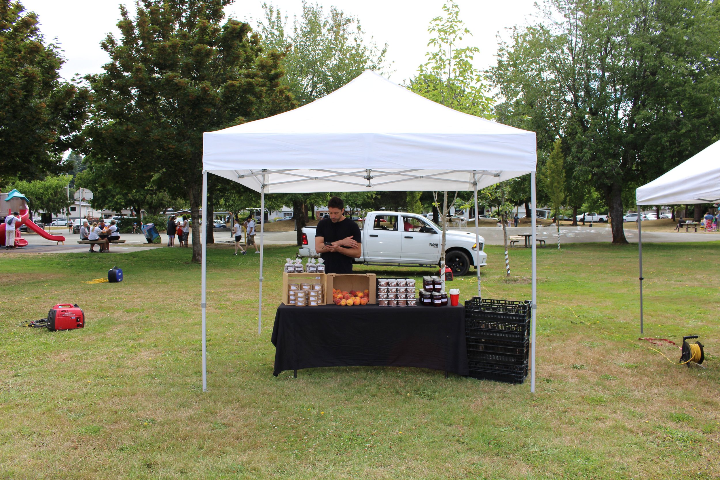 A person standing at a market stall with jars of jam, peaches, and other goods under a white canopy tent in a park.