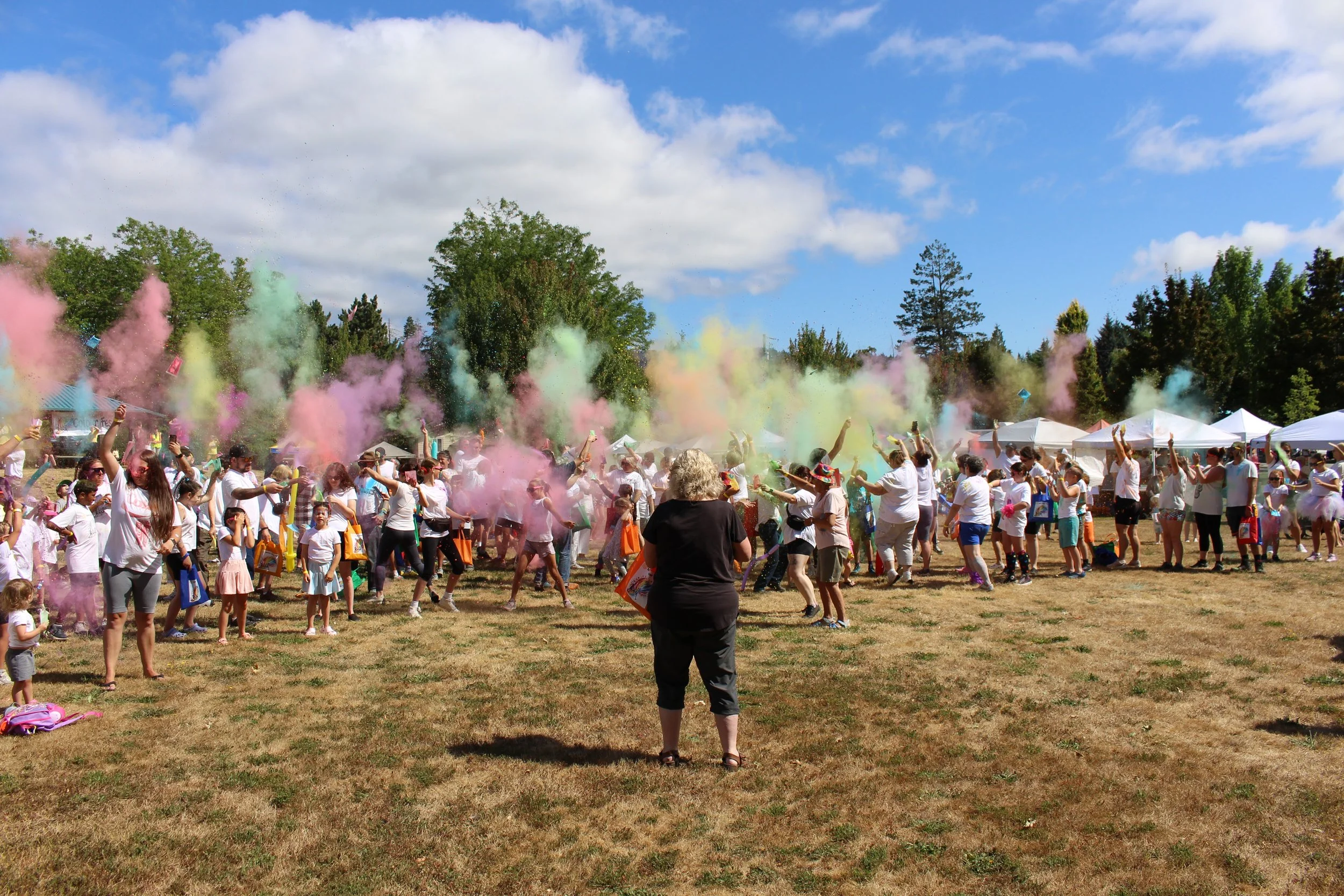People participating in a color powder event outdoors on a sunny day, with colorful clouds of powder in the air and tents in the background.