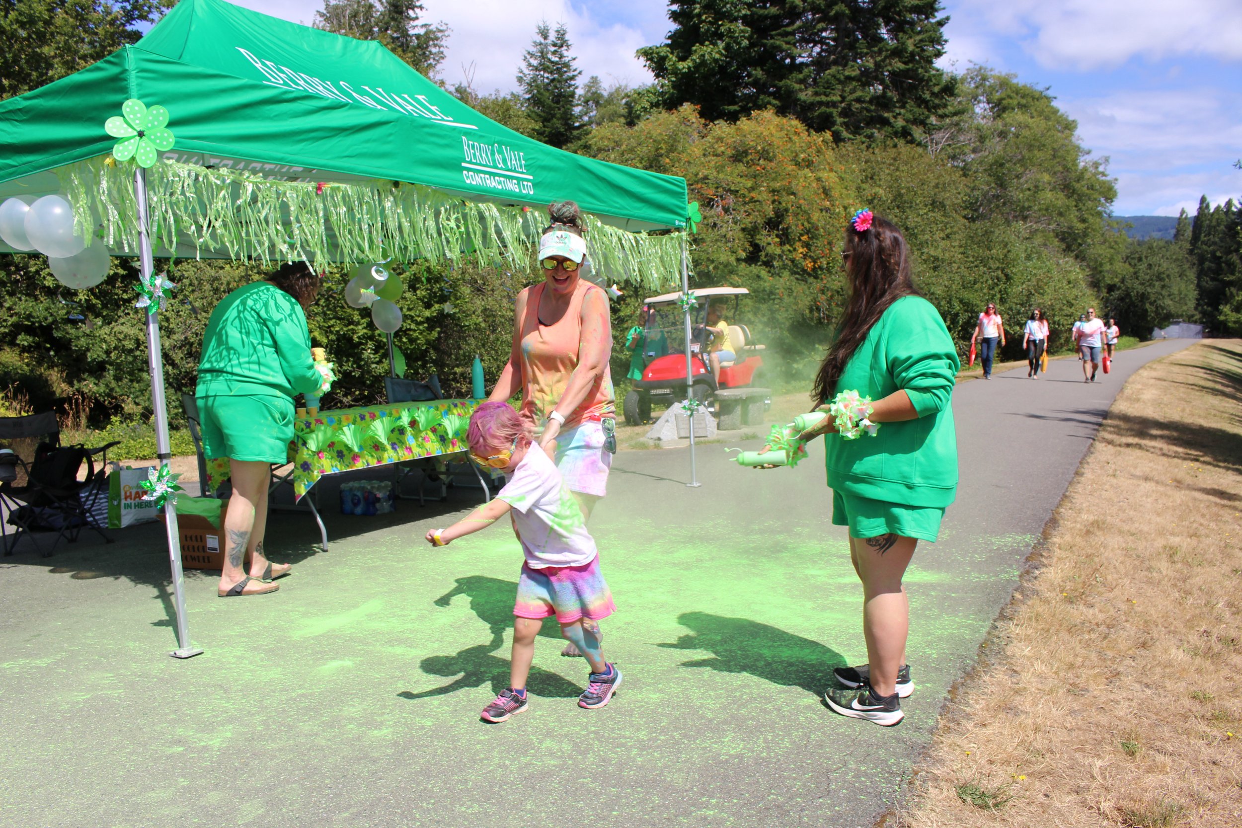 People celebrating a color run event outdoors under a green canopy with green decorations, where a child is being sprinkled with green powder, and others are holding containers with colored powder on a paved pathway surrounded by trees.
