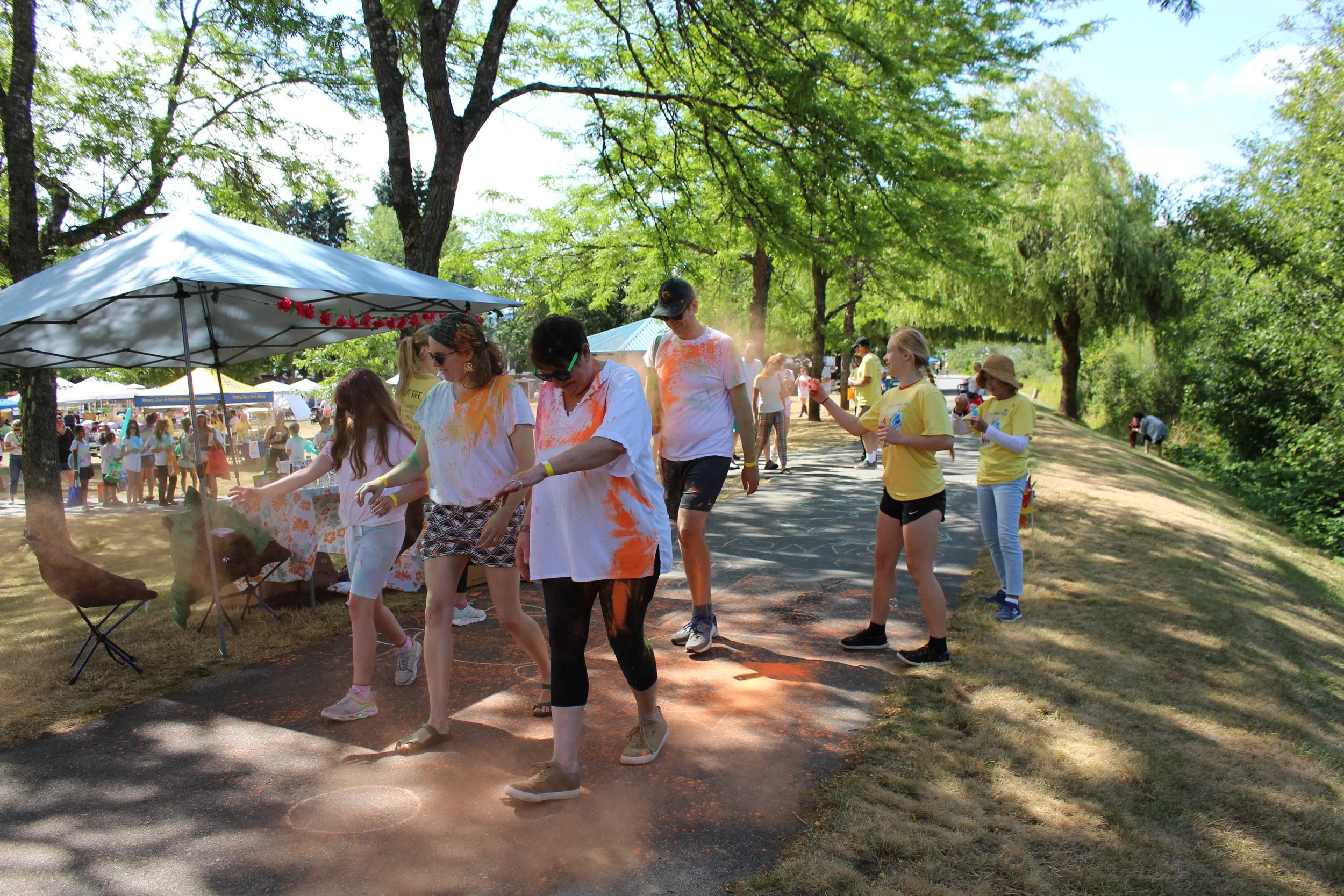 People at an outdoor festival with trees, tents, and a crowd, some wearing colorful splashes of powder.