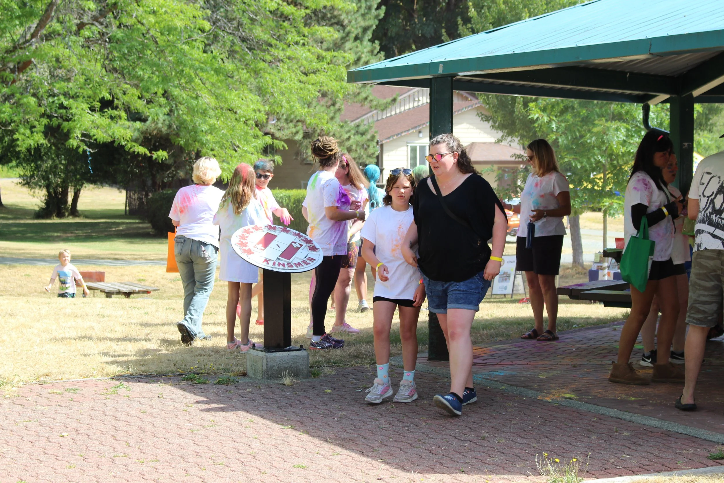 Group of kids and adults participating in a color run event outdoors, with some covered in colorful powder, under a pavilion with green trees in the background.