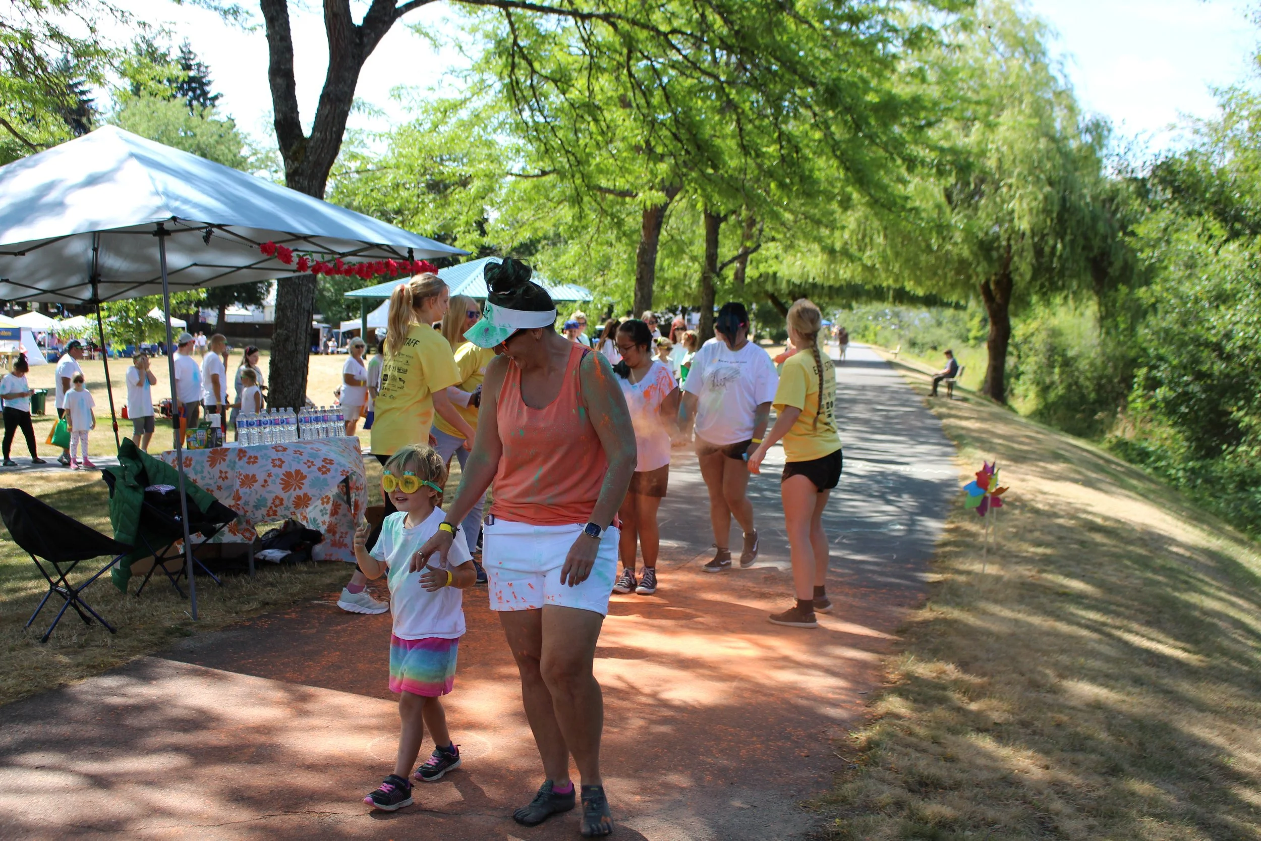 Children and adults at an outdoor summer event, some covered in colorful powder, with tents, trees, and a pathway in a park.