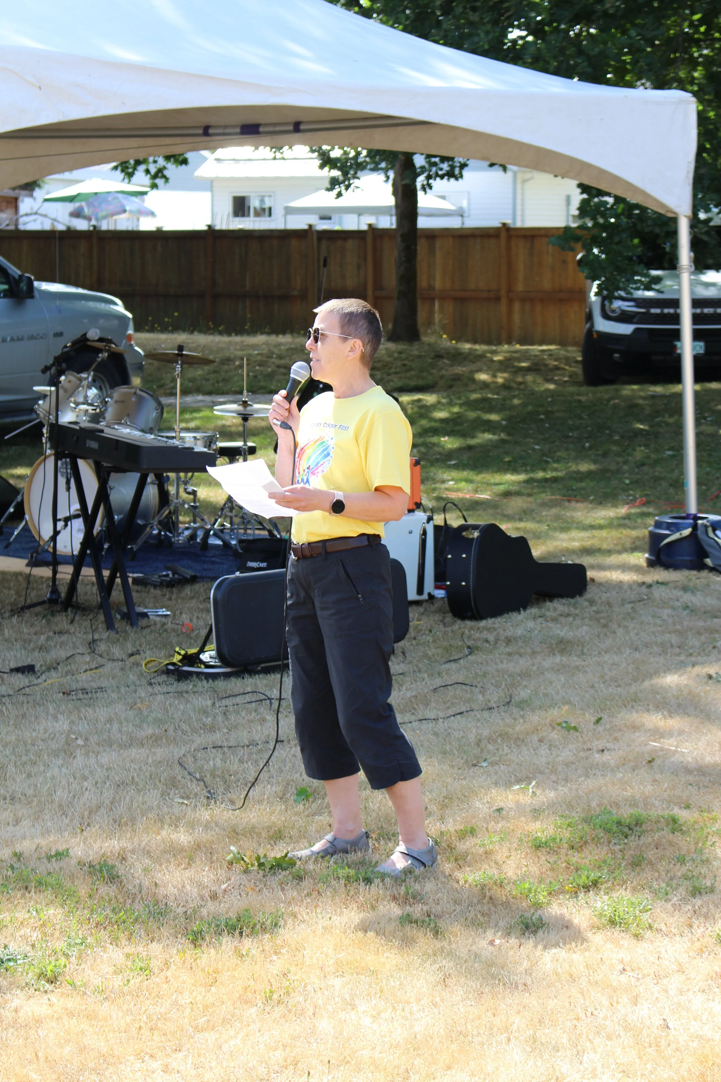 Person holding a microphone and paper, standing outdoors near musical instruments under a white canopy at a sunny event.