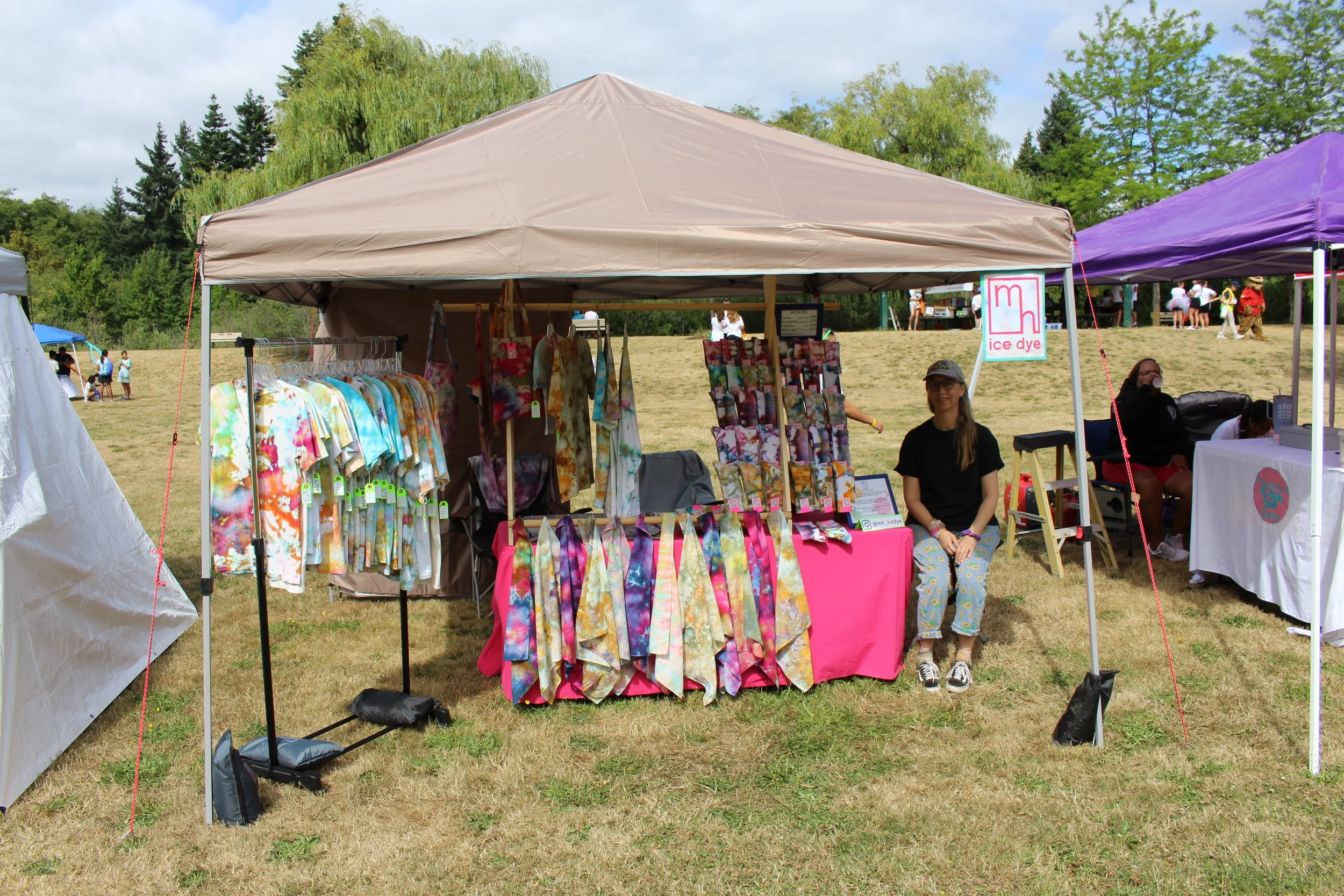 Outdoor market booth selling colorful tie-dye clothing, with a woman sitting at the booth and other vendors nearby, on a grassy area with trees in the background.