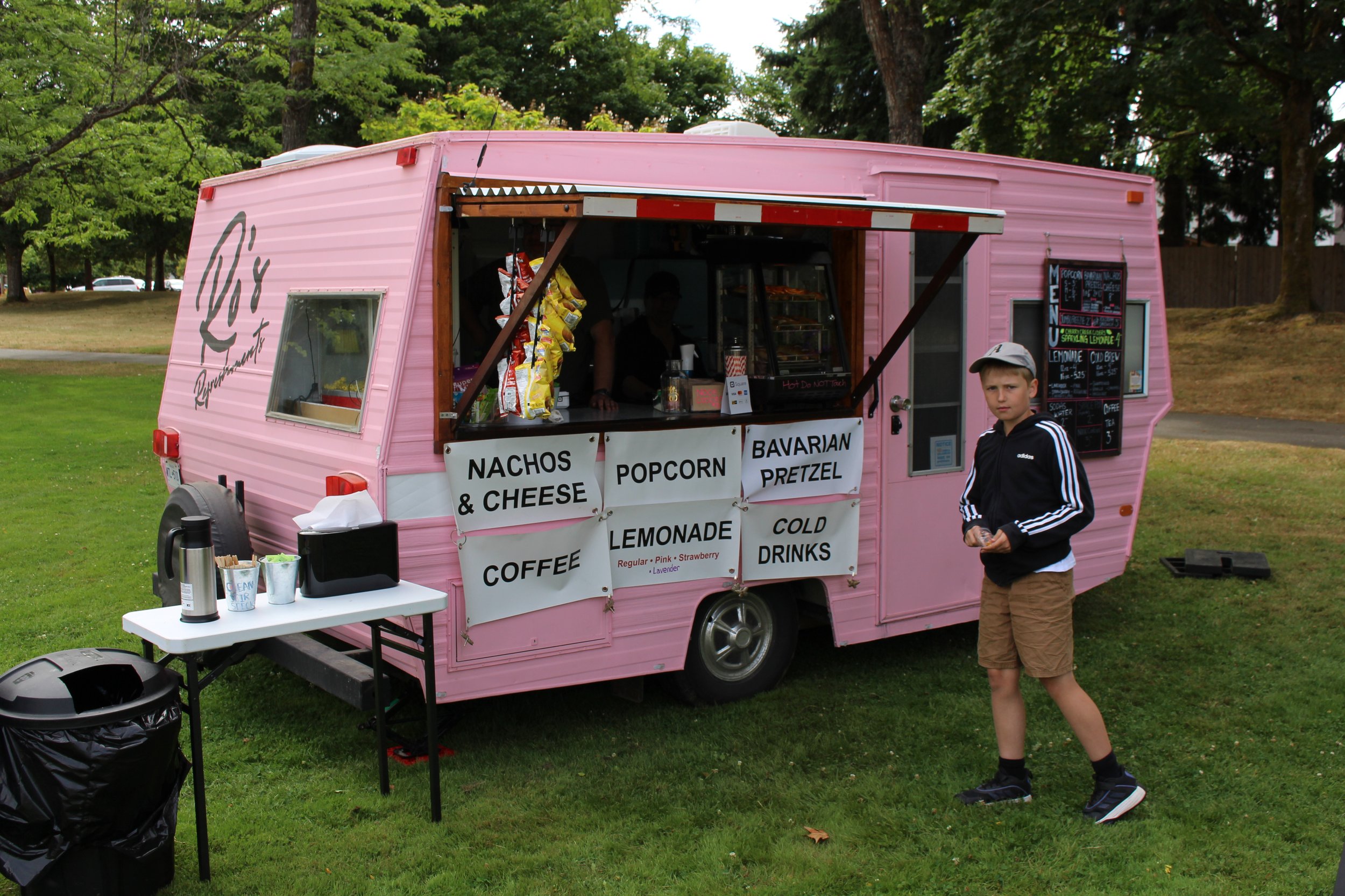 Pink food truck with signs offering nachos and cheese, popcorn, Bavarian pretzel, coffee, lemonade, and cold drinks, with a boy standing in front of it on grass in a park.