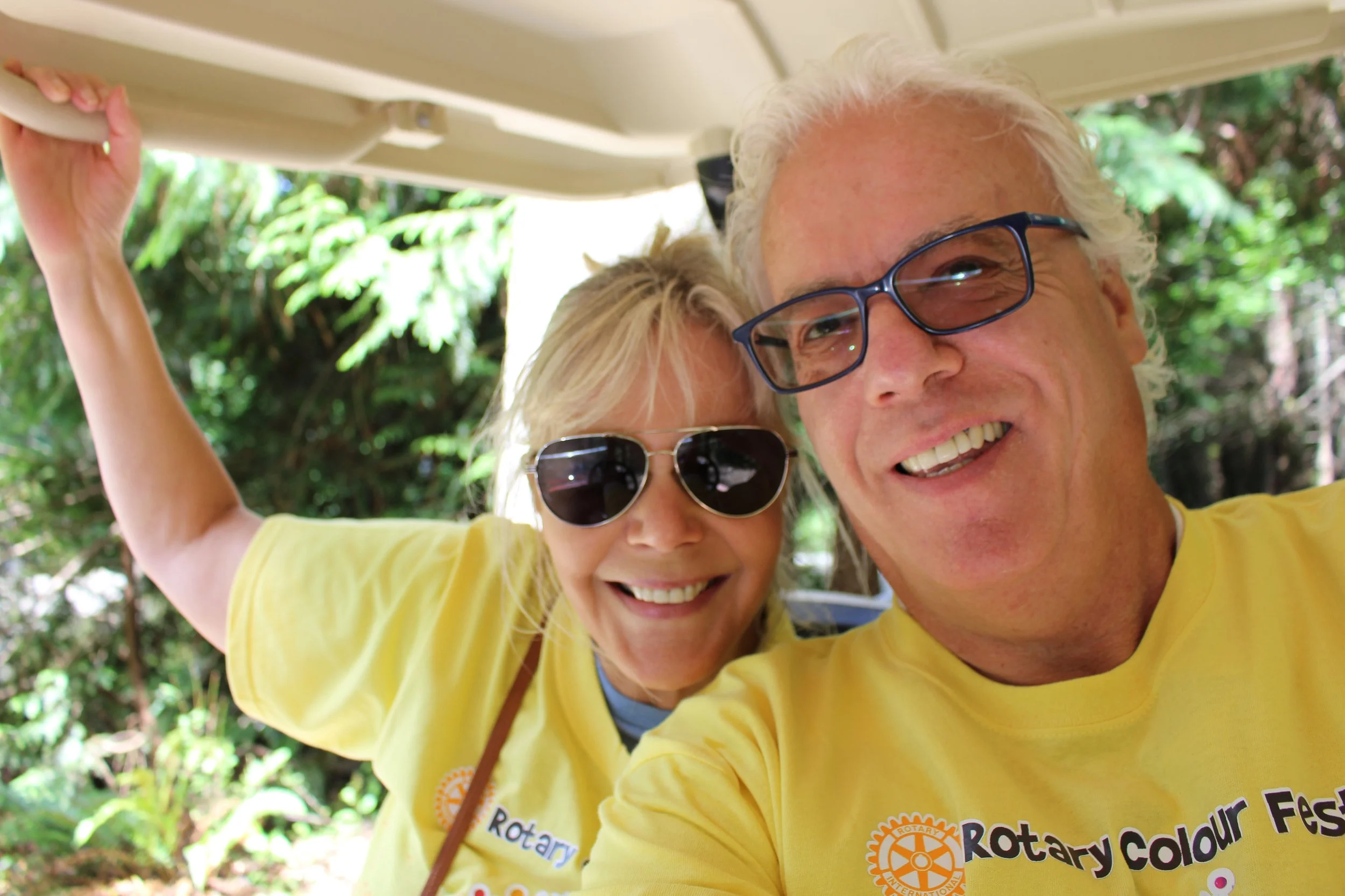 Smiling elderly man and woman wearing yellow Rotary shirts taking a selfie outdoors in a green, wooded area. The man is wearing glasses and the woman sunglasses, both with happy expressions.
