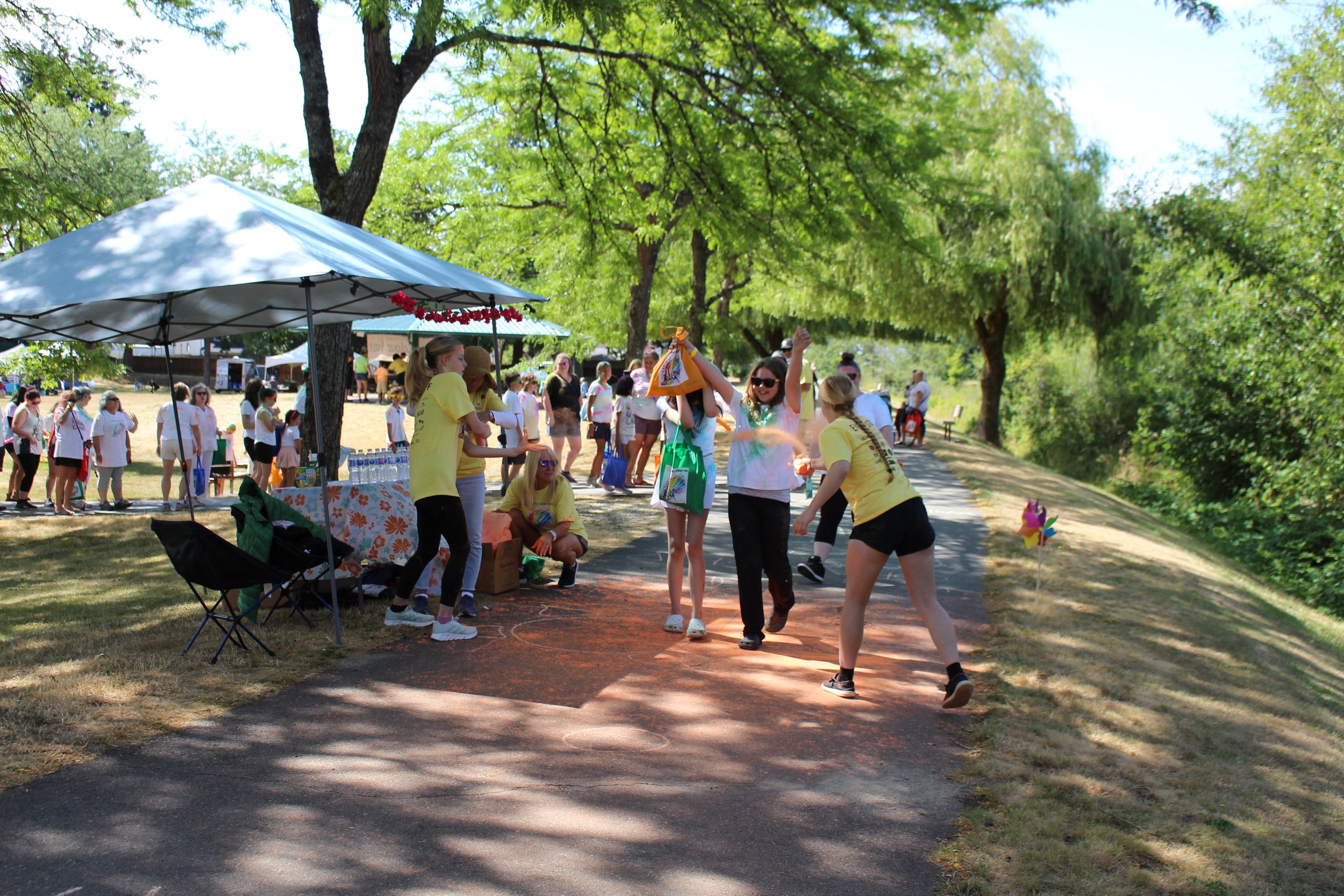 People enjoying a festival outdoors on a sunny day with a group dancing in the center, a water-themed game, and booths under a large umbrella, surrounded by green trees and a pathway.