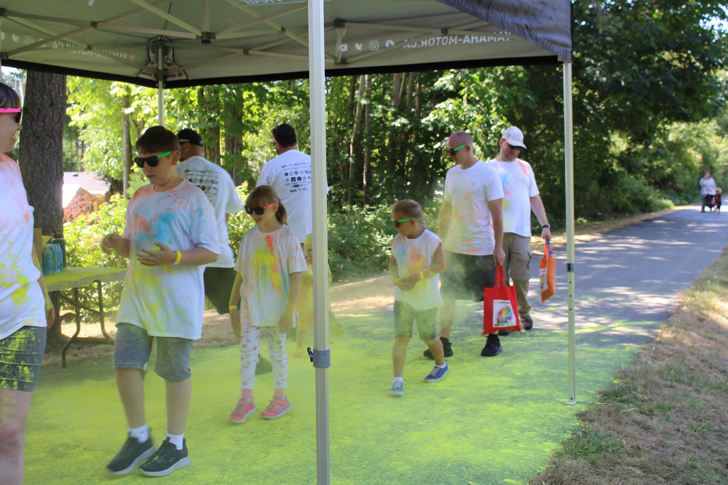 People participating in a color run event, walking through a yellow powder cloud under a green canopy in a park, with trees and a walking path in the background.