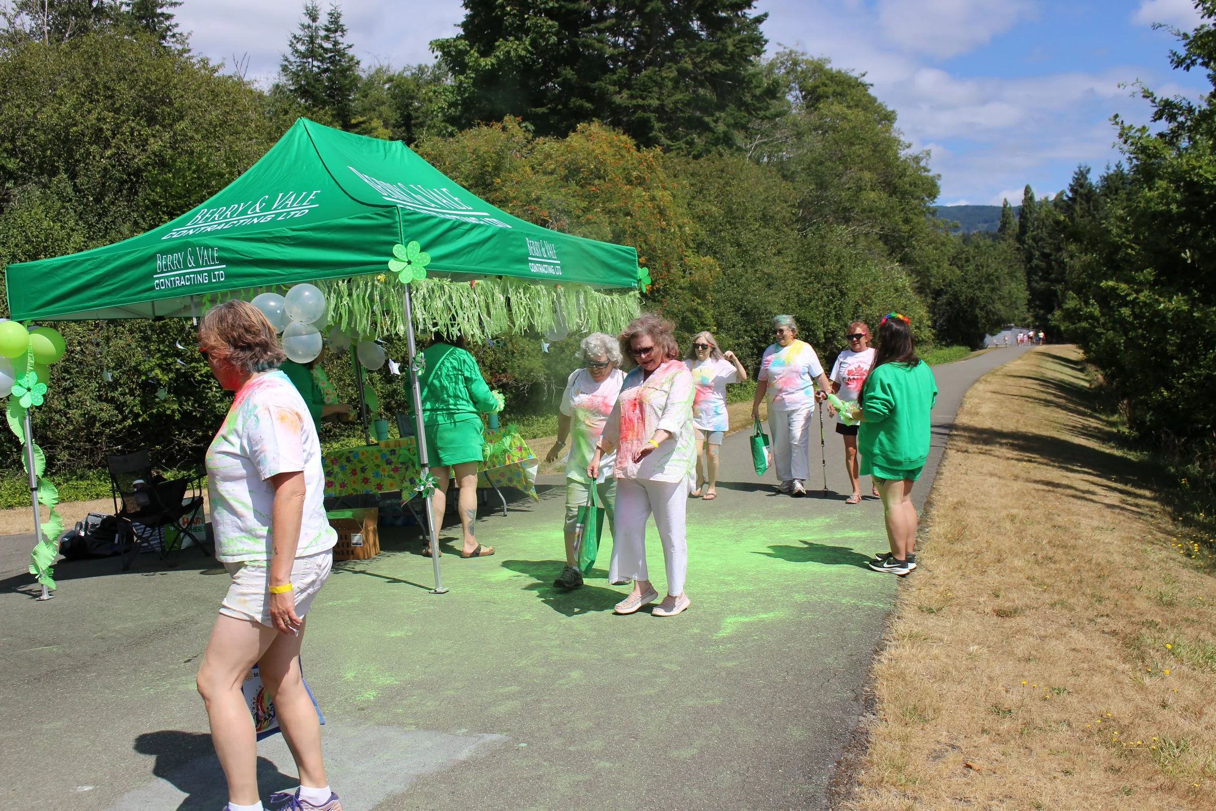 People participating in a color run, with their white clothes covered in colorful powder, standing near a green tent decorated with shamrocks, on a sunny day along a tree-lined path.