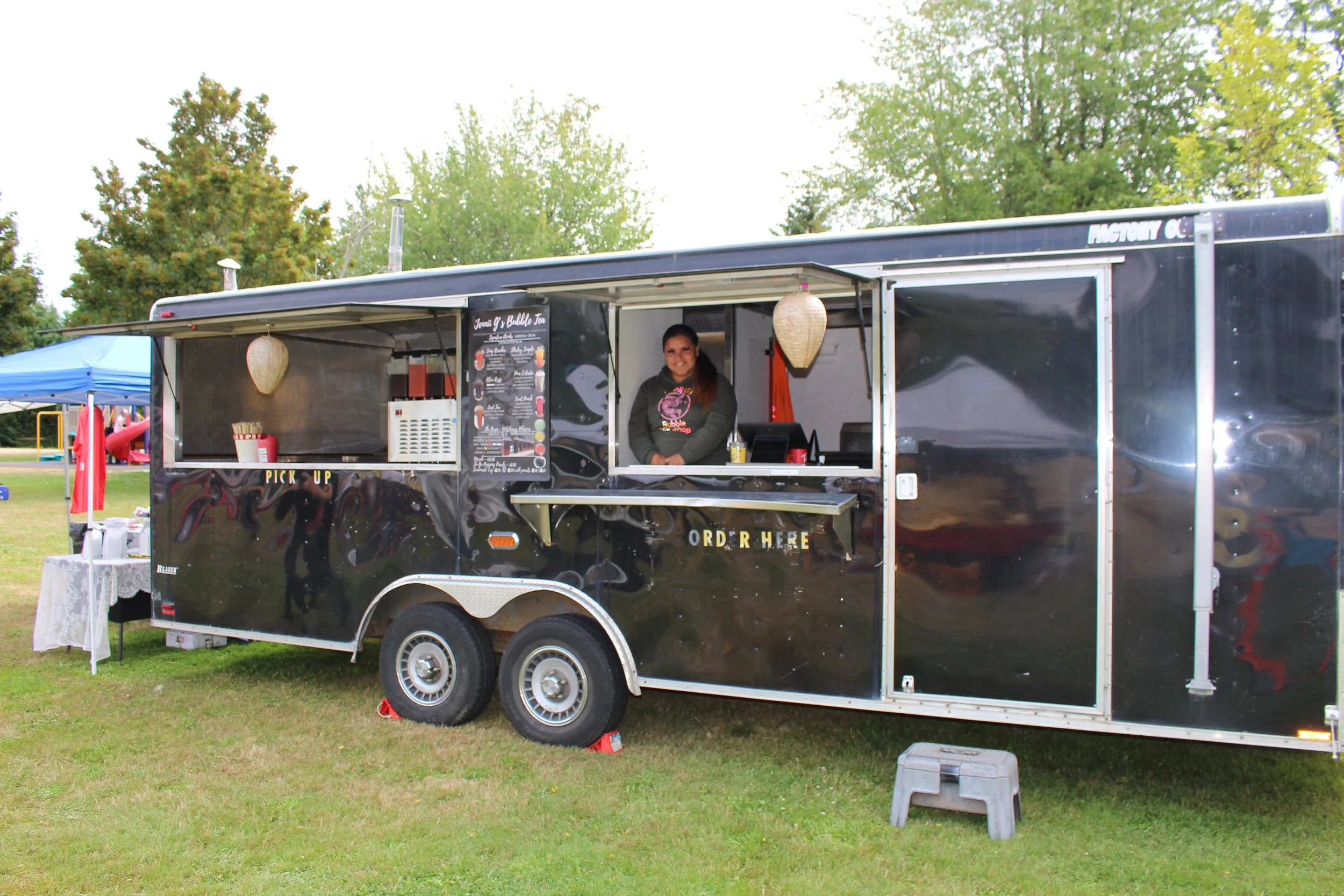 Black food truck with a woman inside, serving at a park with green trees, a blue tent, and playground equipment in the background.