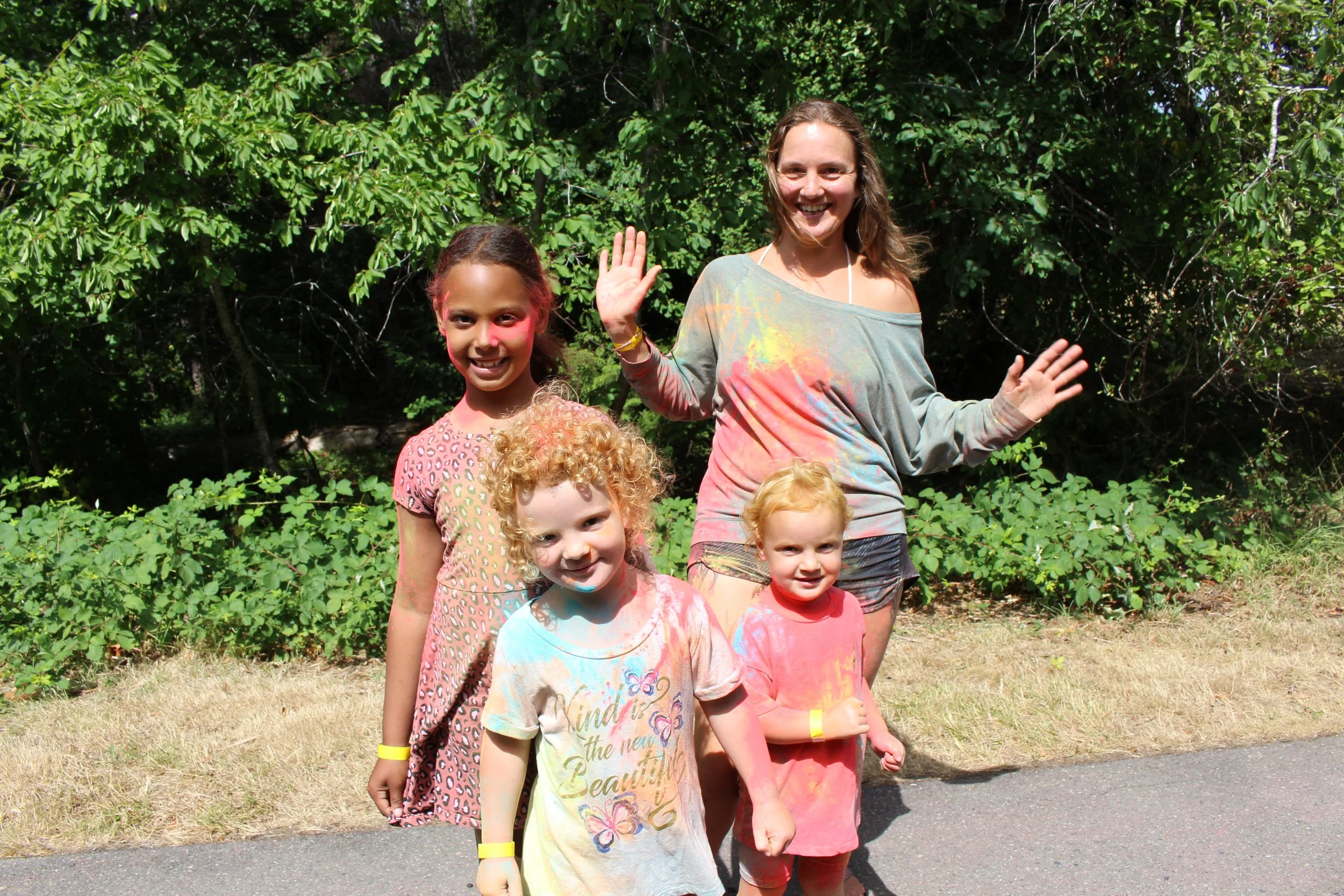 A group of four children and one adult woman celebrating a color festival outdoors, with colorful powder on their faces and clothes, standing on a paved path surrounded by greenery.