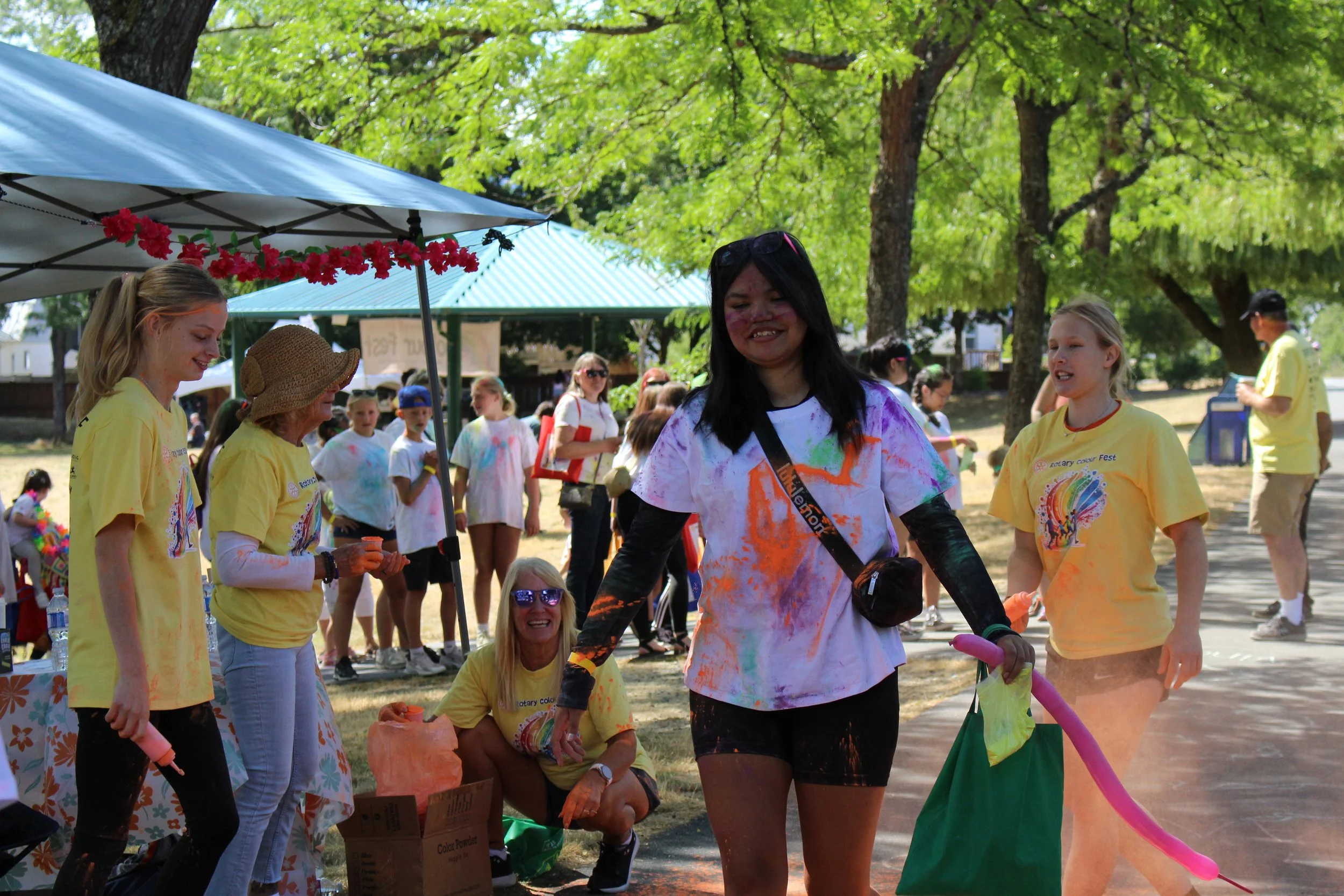 People celebrating at a colorful outdoor event, covered in vibrant powder during the color run, with trees and a tent in the background.