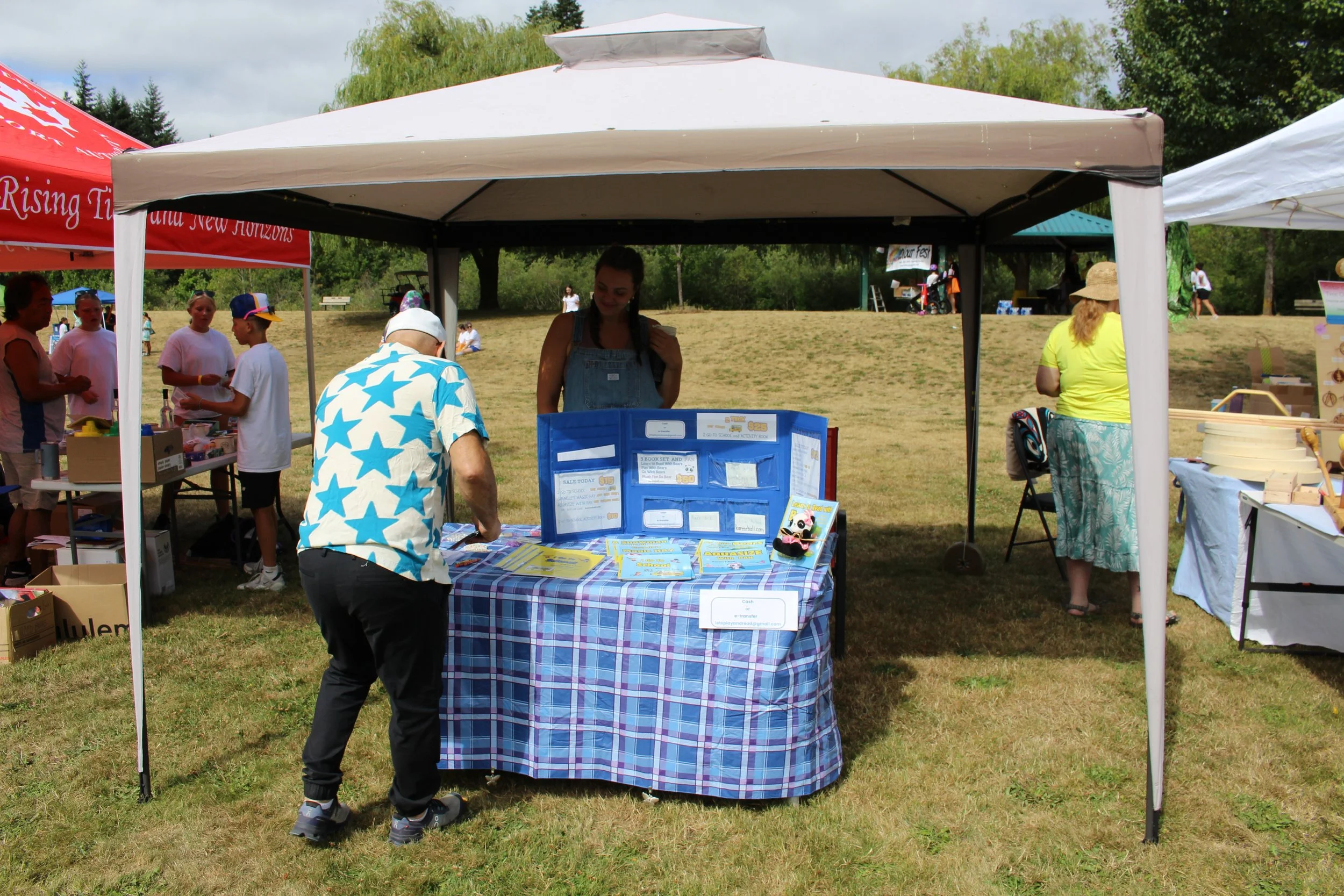 A scene at an outdoor fair with a booth under a white canopy. A woman behind the booth interacts with a man in a star-patterned shirt. Other booths and people are visible in the background on a grassy area with trees.
