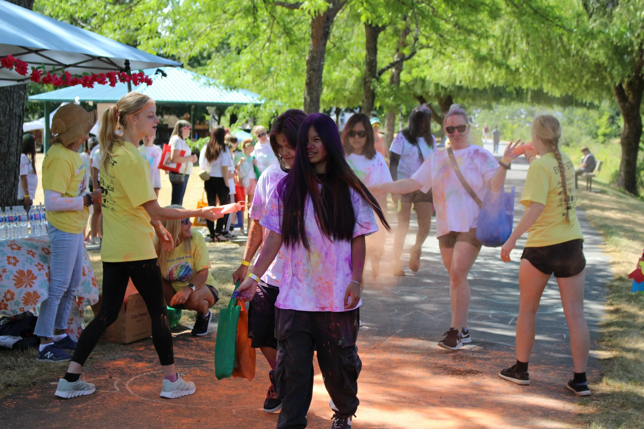 People participating in a color run, with colorful powder on their clothes and skin, on a sunny day under green trees, with tents and other participants in the background.