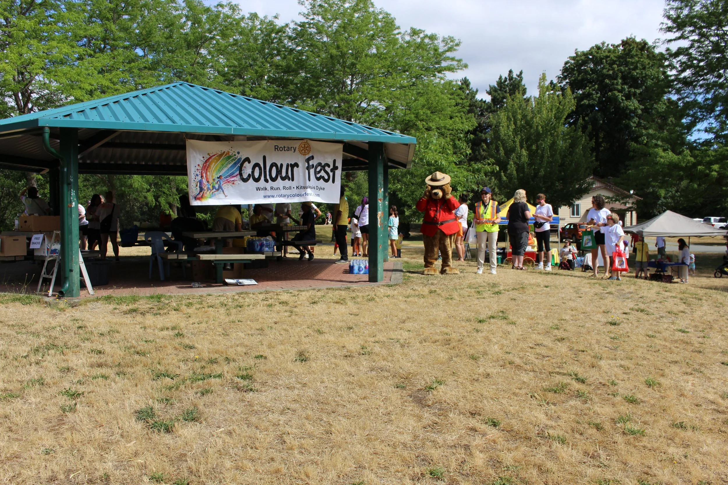 Community event in a park with a banner reading 'Colour Fest,' people gathered around tables, a person in a bear mascot costume, and a white tent, with green trees in the background.
