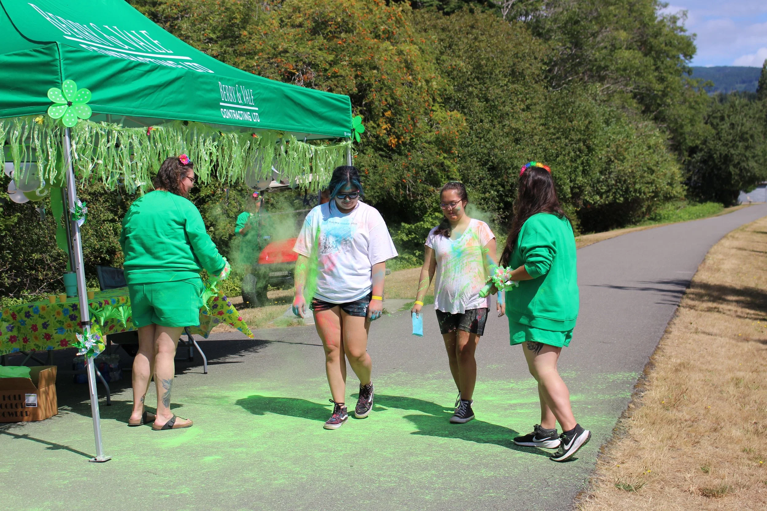 Three girls and two women participating in a color run, covered in colored powder, near a green canopy with St. Patrick's Day decorations outdoors on a sunny day.