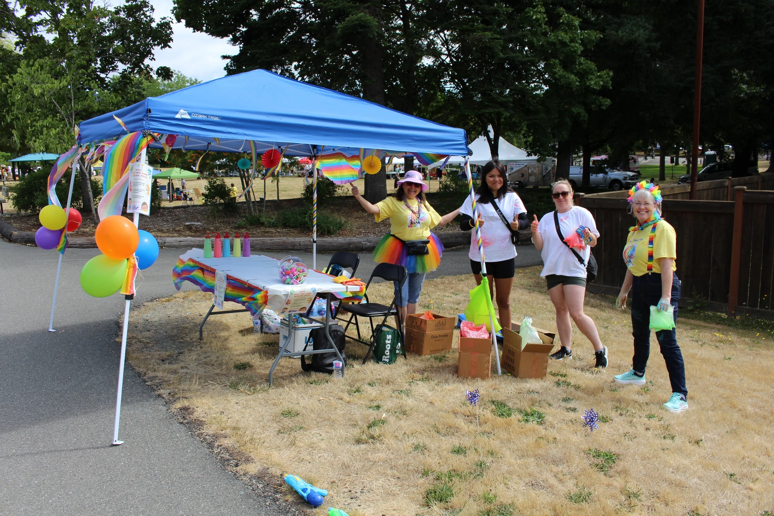 Four women standing at a rainbow-themed outdoor event booth with balloons, colorful decorations, and supplies under a blue canopy tent, with trees and other booths in the background.