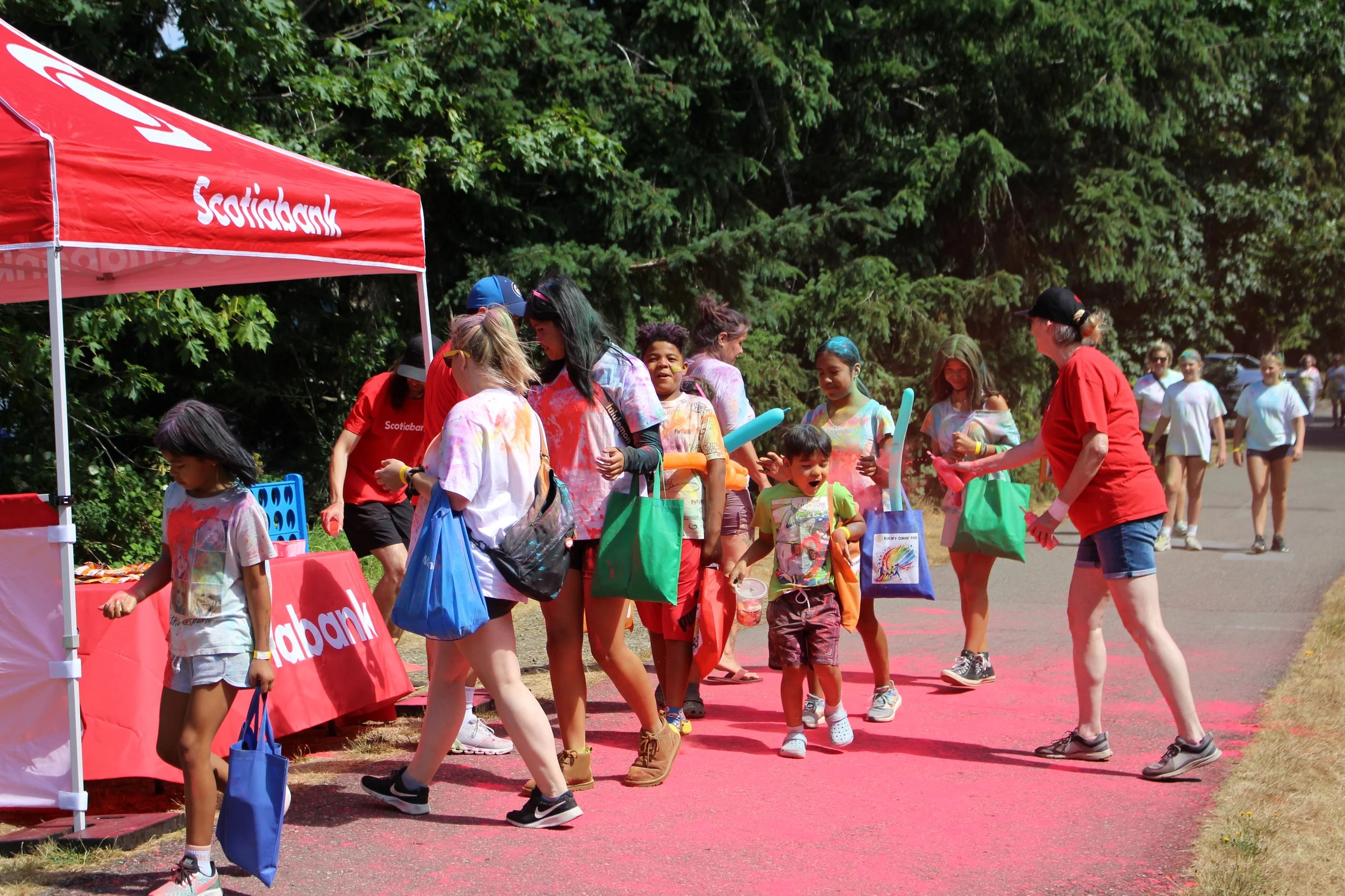 Children and adults gathering around a red Scozipic tent at a color run event, with colorful powder on their shirts and on the ground.