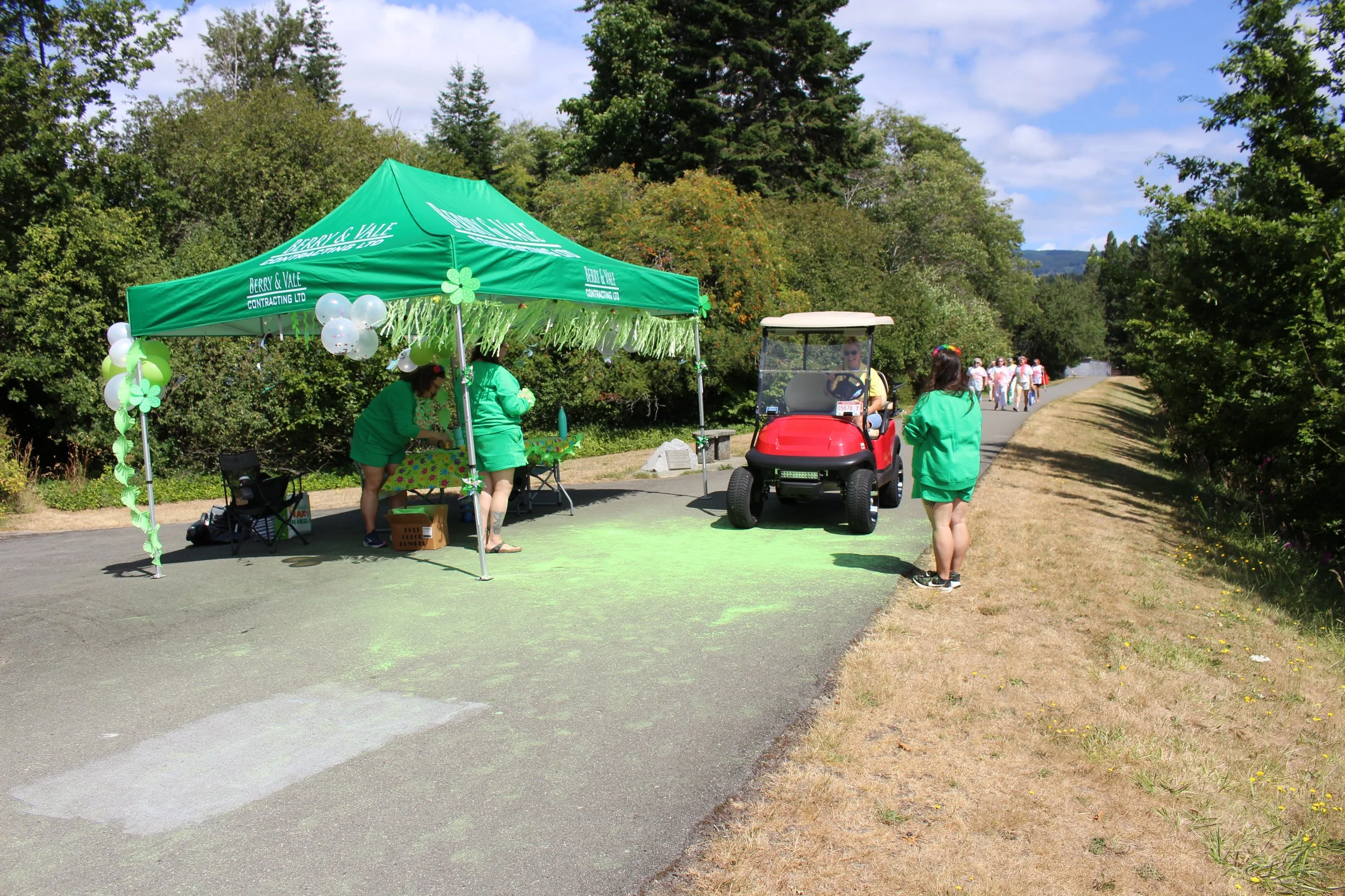 People at a festive outdoor event on a paved path with a green tent decorated with balloons and shamrocks, a woman in a yellow shirt driving a golf cart, and a woman in green standing nearby during daylight.