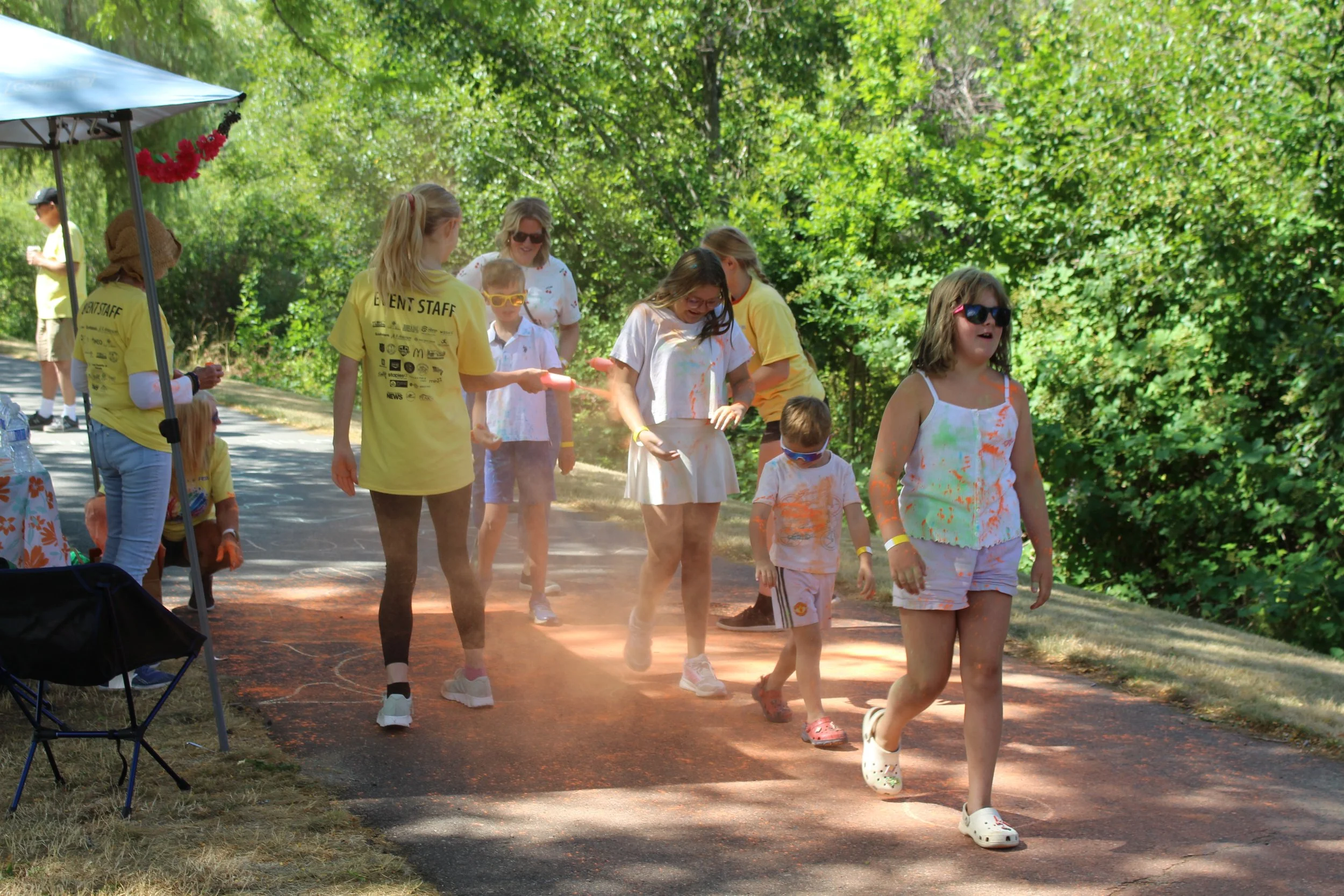 Kids and adults participating in a color run event outdoors, wearing white shirts splattered with colorful powder, on a paved pathway surrounded by green trees.