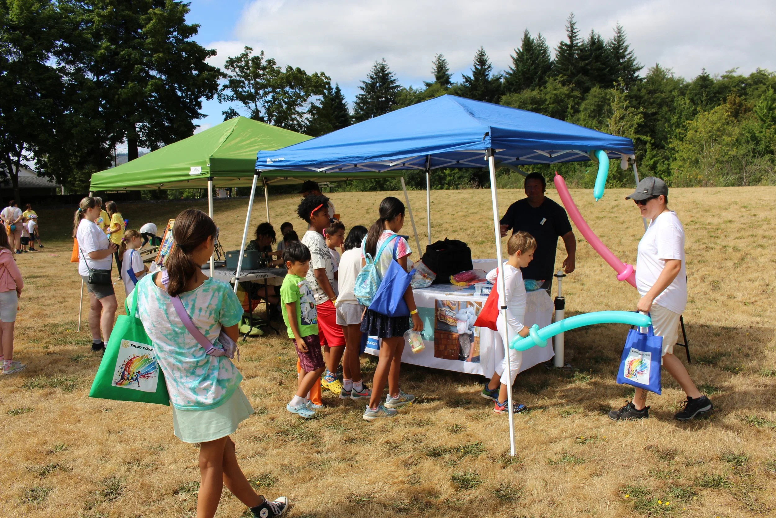 Children and adults lined up at a tented outdoor event, waiting for food or activities, with balloon twisting by two volunteers.