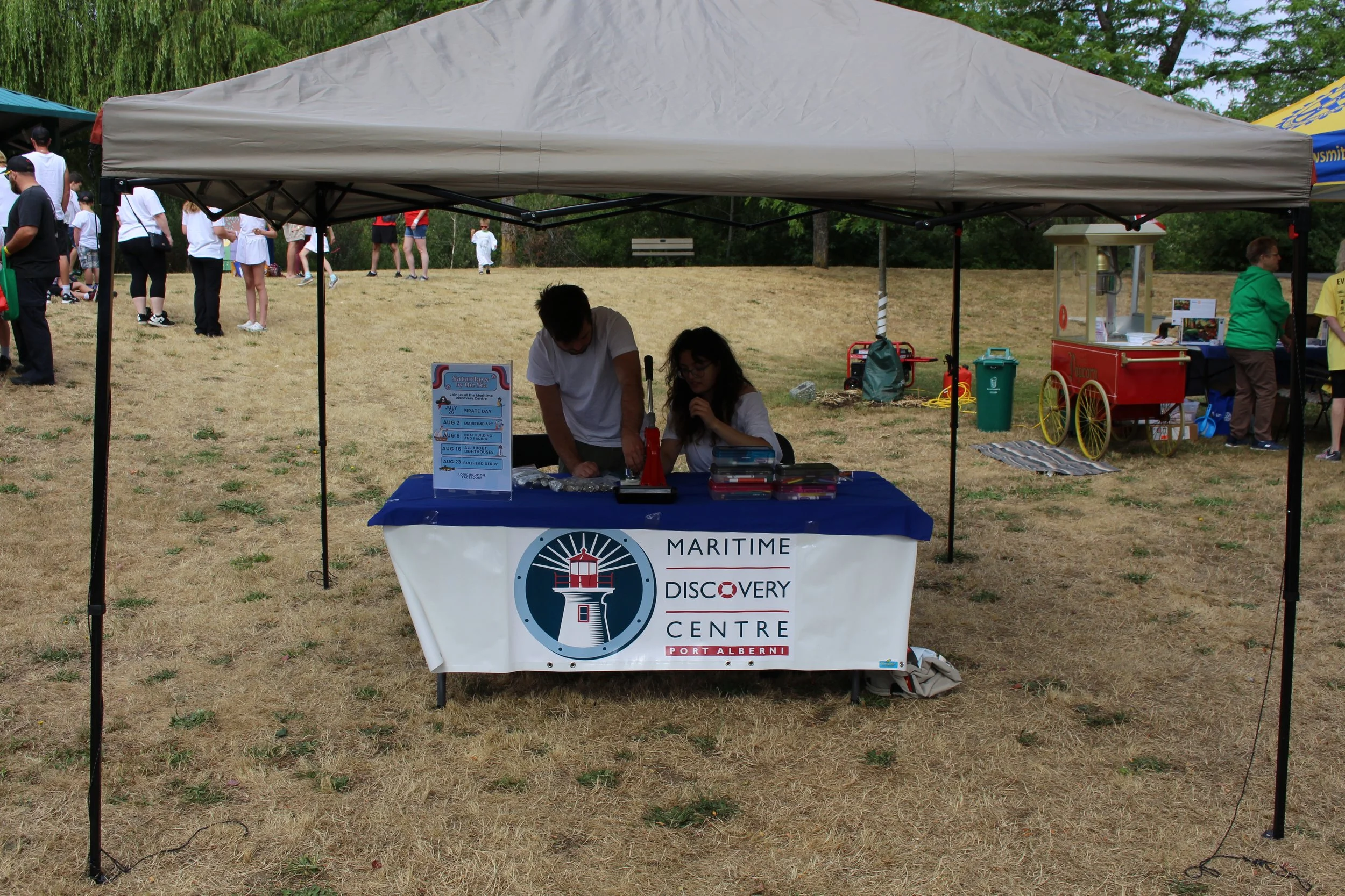A tent at an outdoor event with a sign that reads "Maritime Discovery Centre", showing two people working with some items on a table, and a group of people in the background.