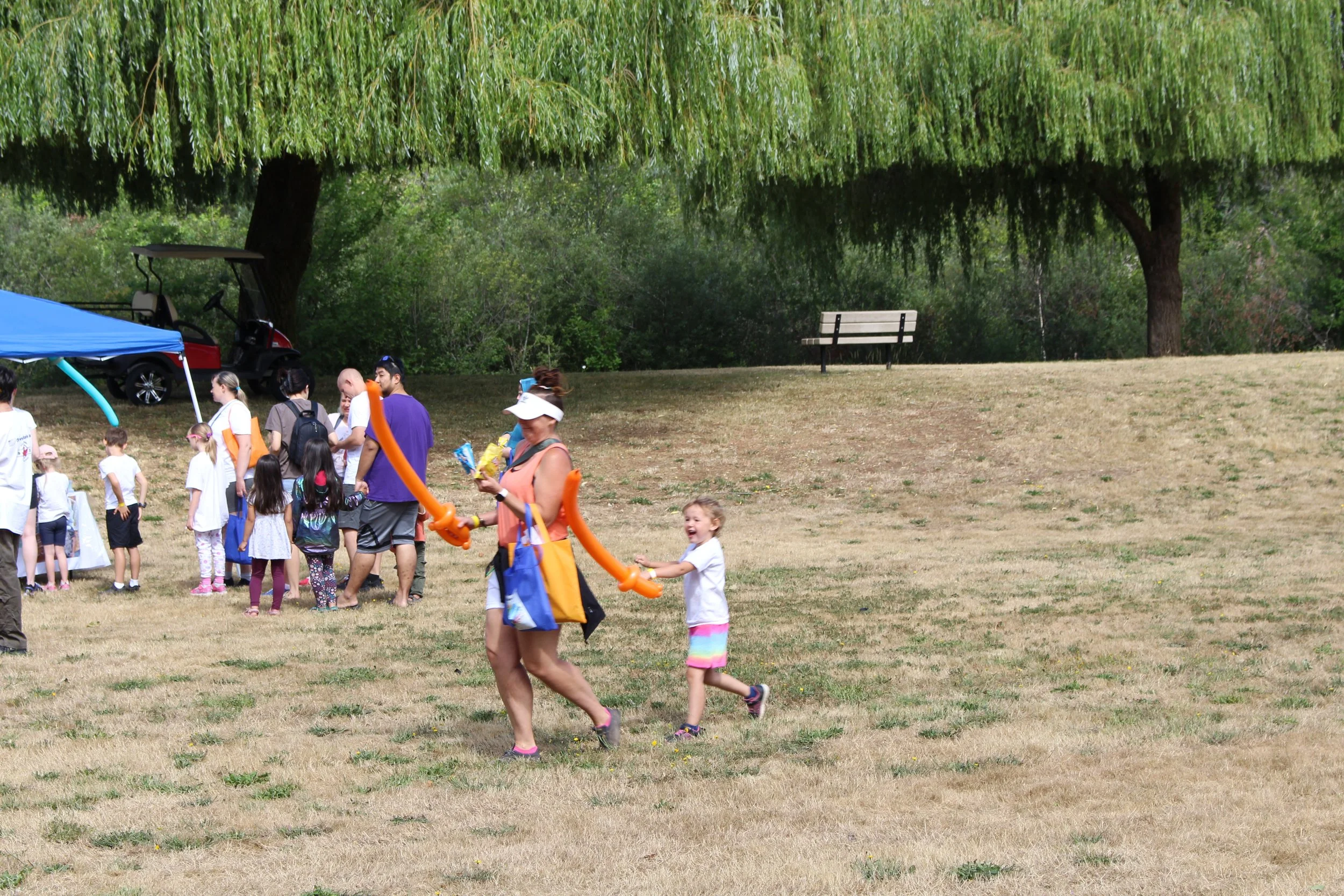 Children and adults at an outdoor event on a sunny day, some children playing with balloon animals, a line of people near a table, trees and a park bench in the background.