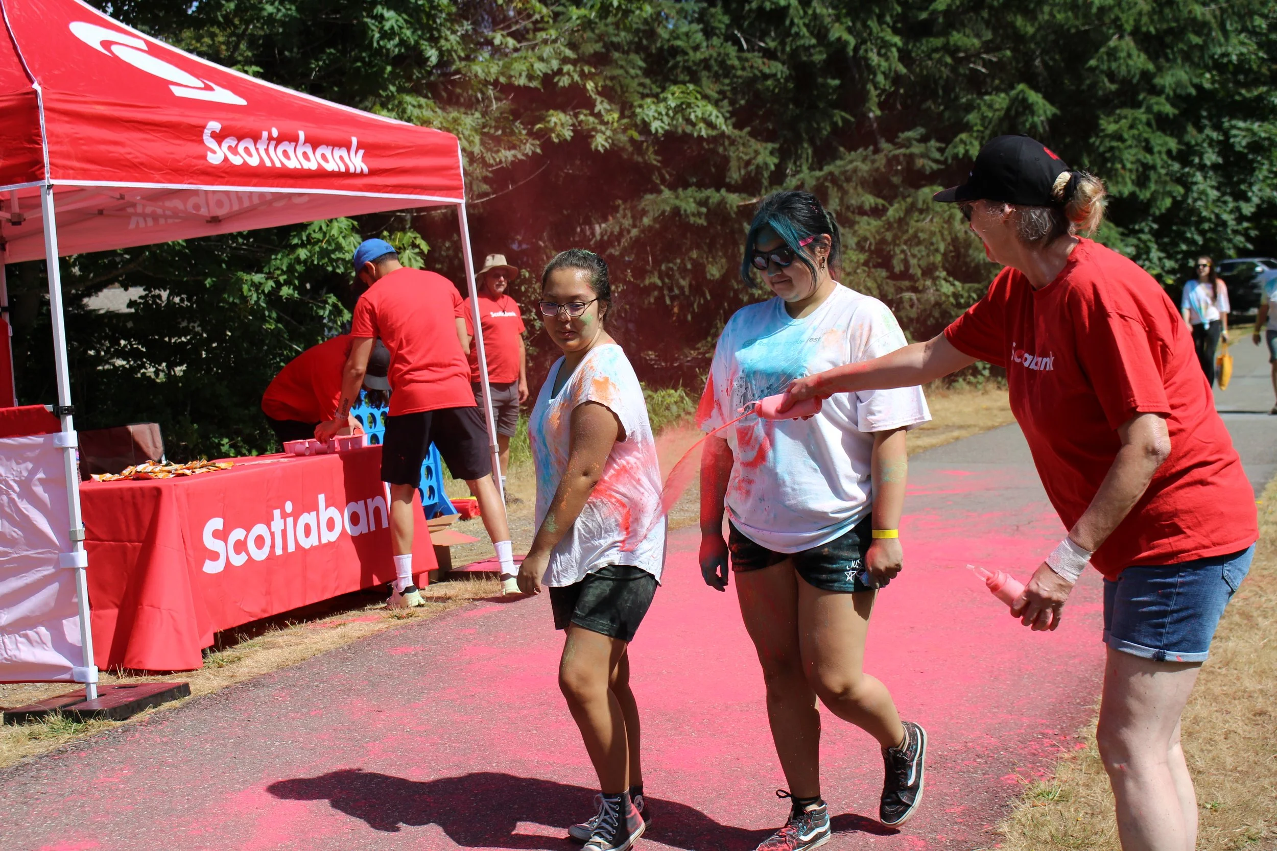 Three women covered in pink and orange powder walking during a color run event near a red Scotiabank tent, with other participants and trees in the background.